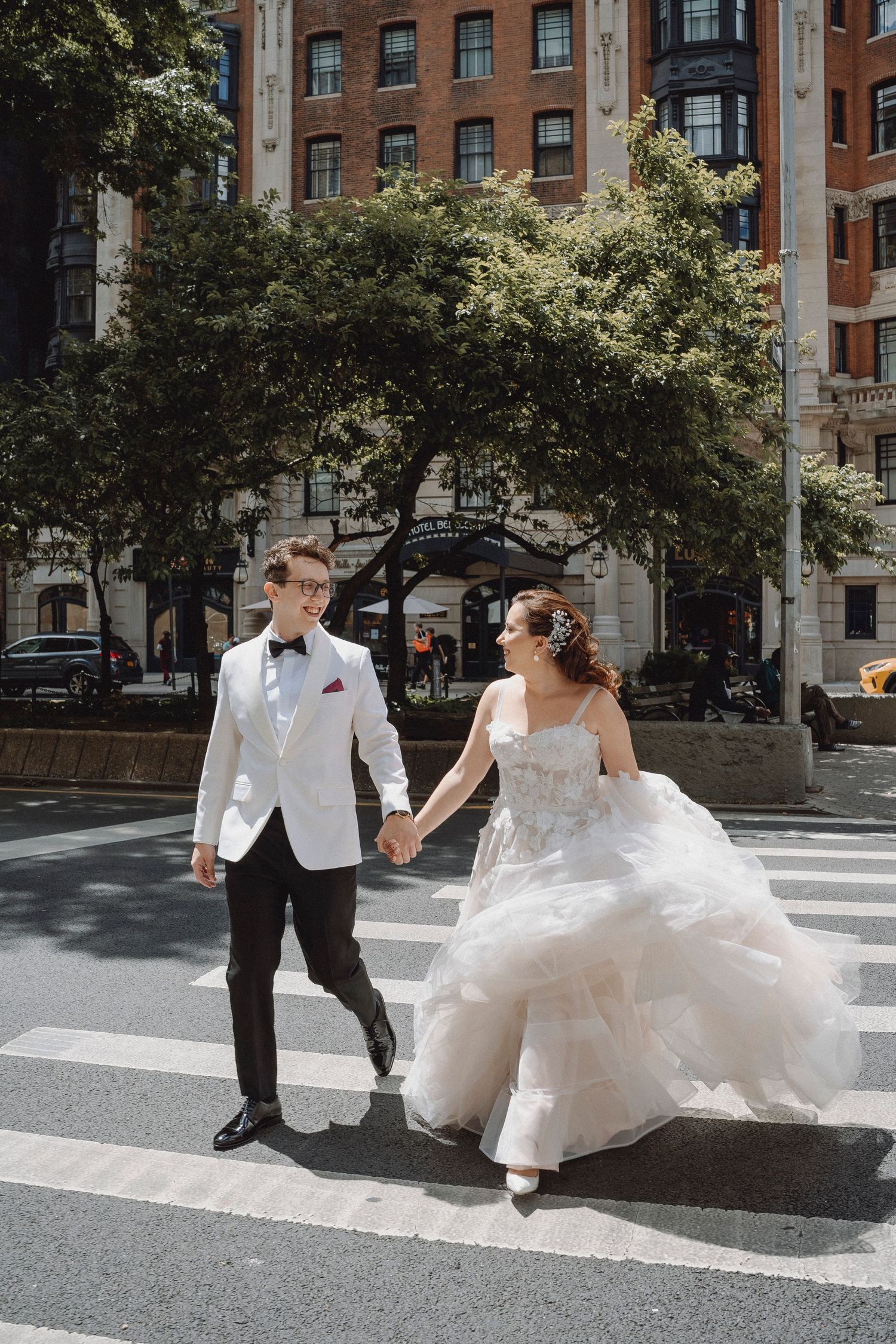 Bride in tulle gown and groom in white tuxedo cross city street holding hands.