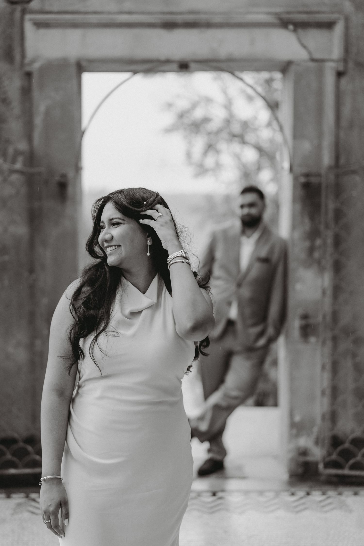 Black and white photo of smiling bride in white dress with groom standing in archway background.