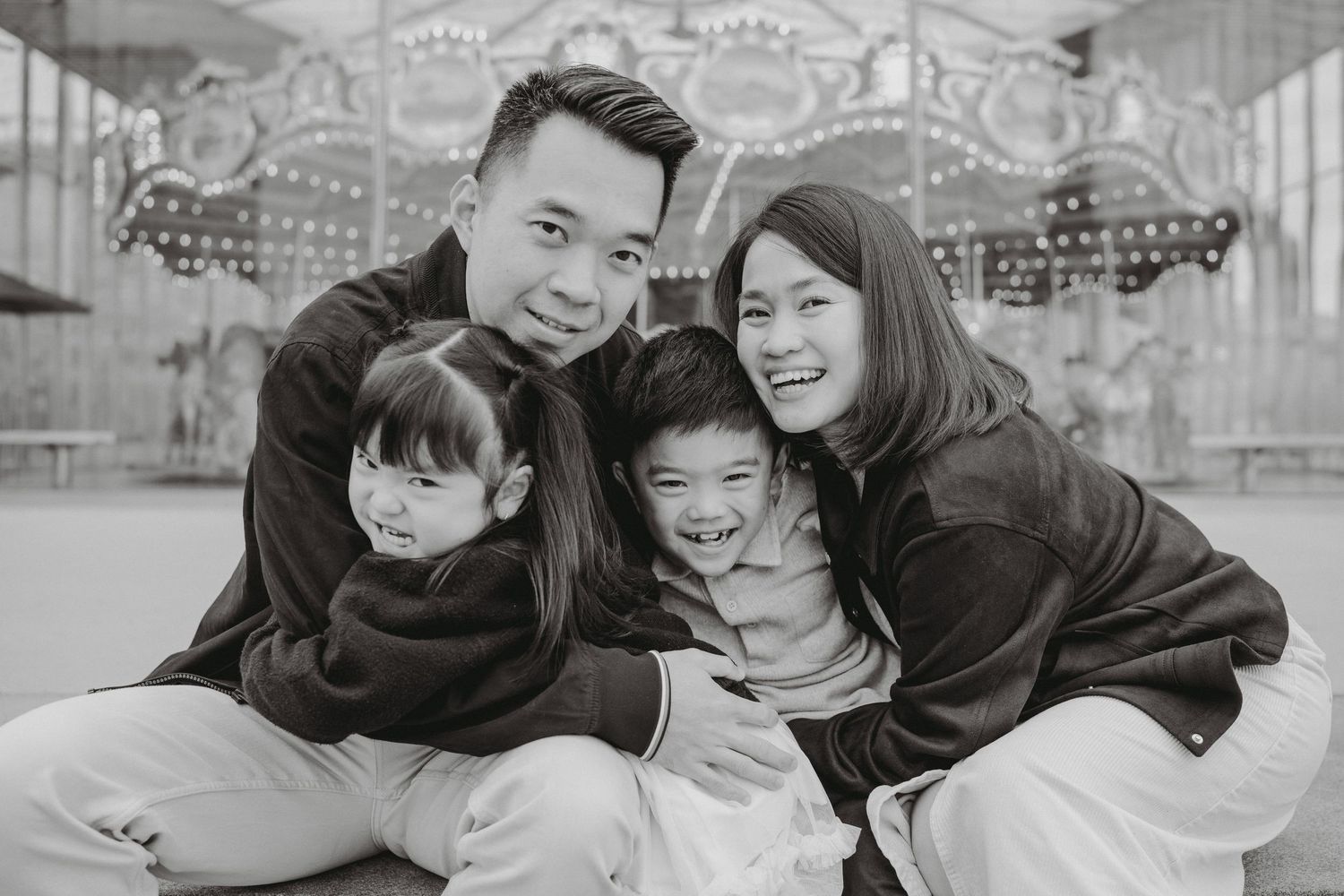 Happy family of four poses together in black and white photo in front of a carousel.