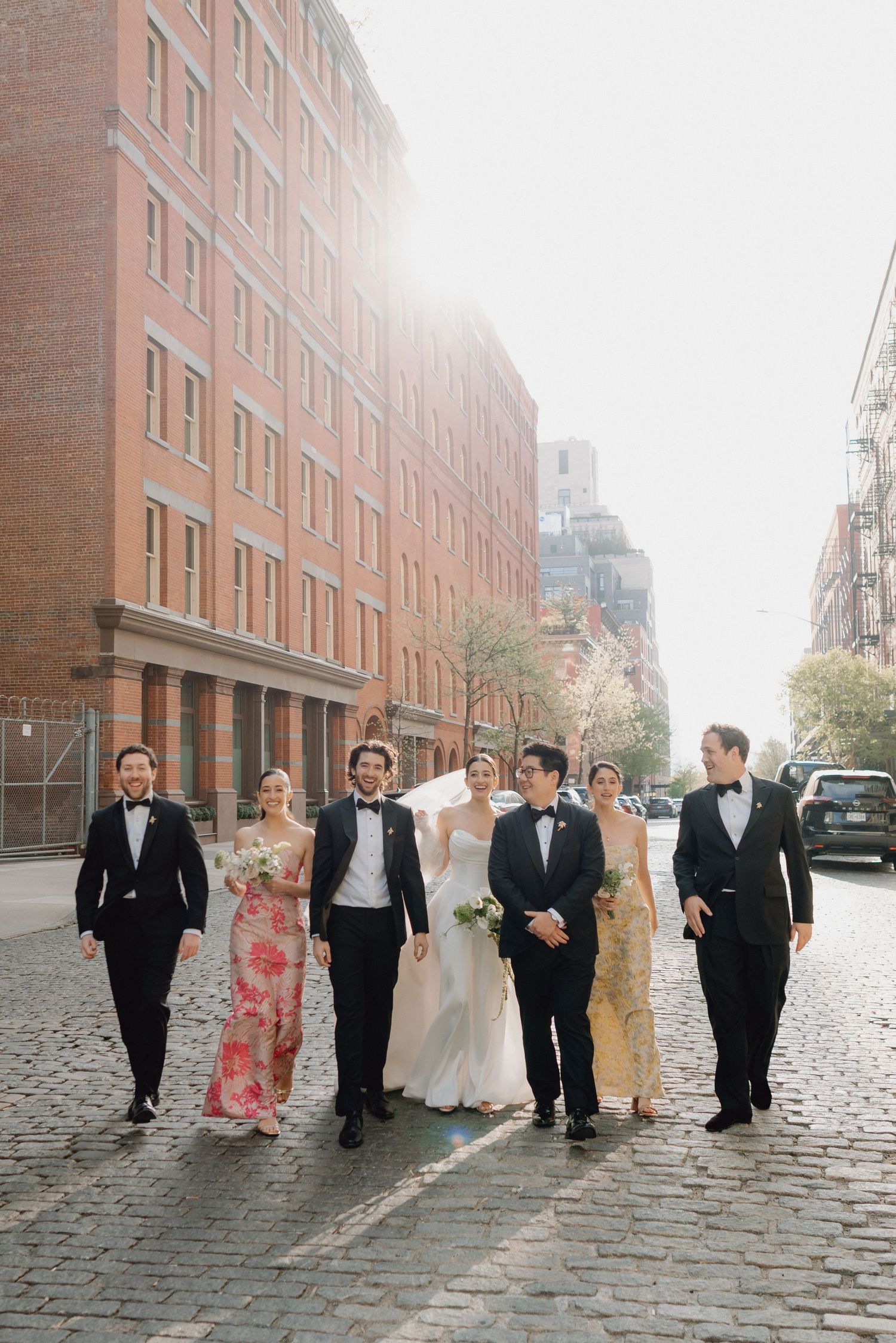 Wedding party walks together down sunny cobblestone street in New York City neighborhood.