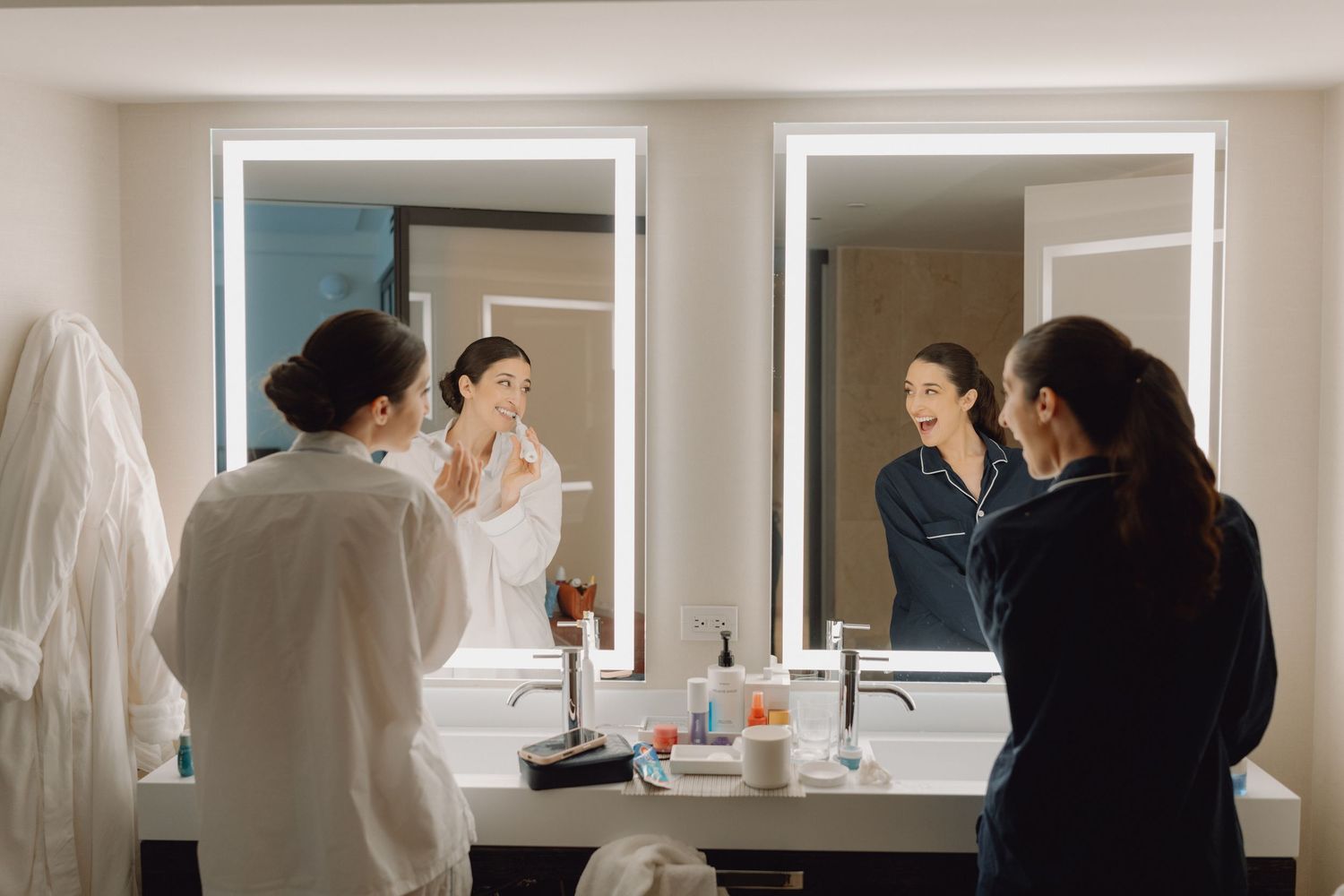 Bride and bridesmaid laugh together while getting ready in a modern hotel bathroom with lit mirrors.