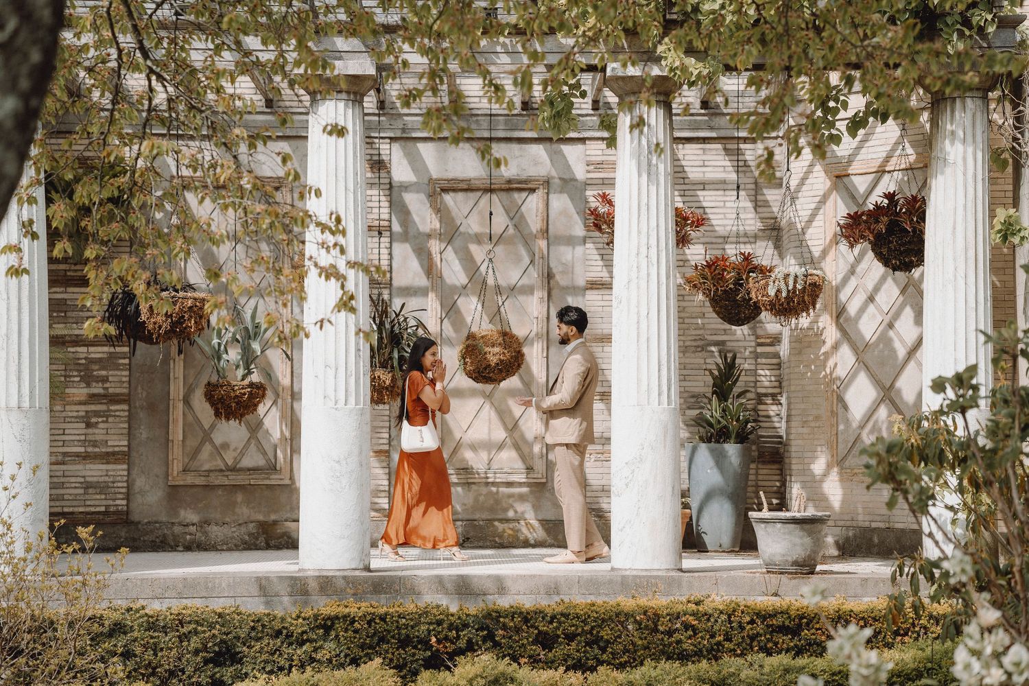 Couple in rust and beige outfits stand between white columns of a sunlit garden pergola.