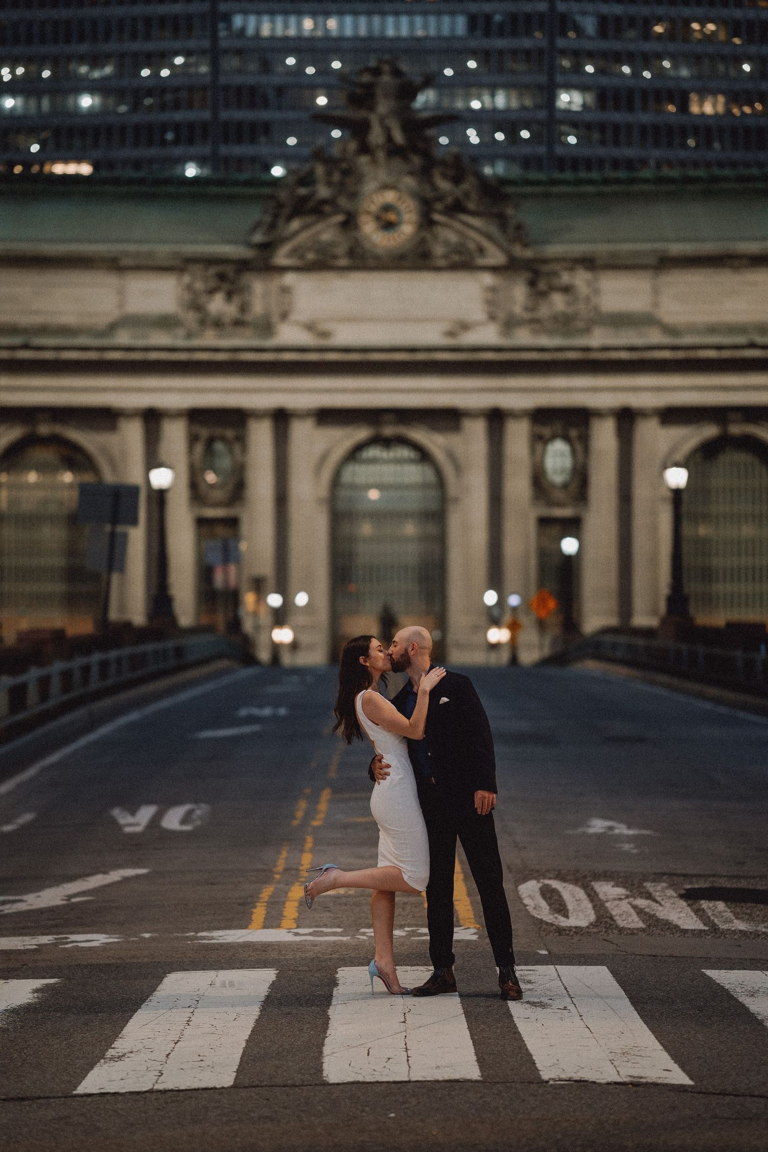 Couple kissing on a NYC crosswalk at night with Grand Central Terminal facade behind them.