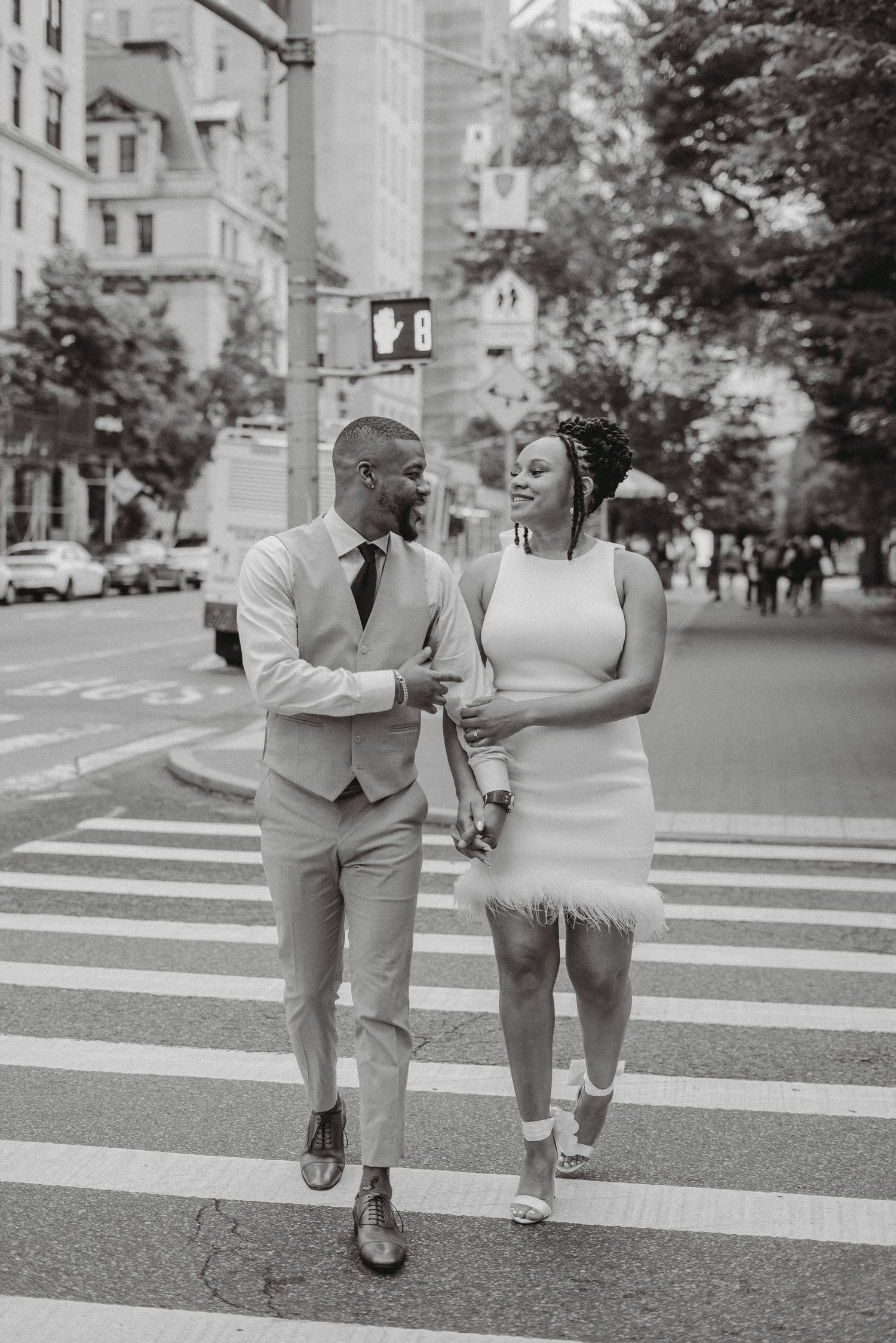 Black and white photo of an elegant couple crossing a city street, smiling at each other in wedding attire.