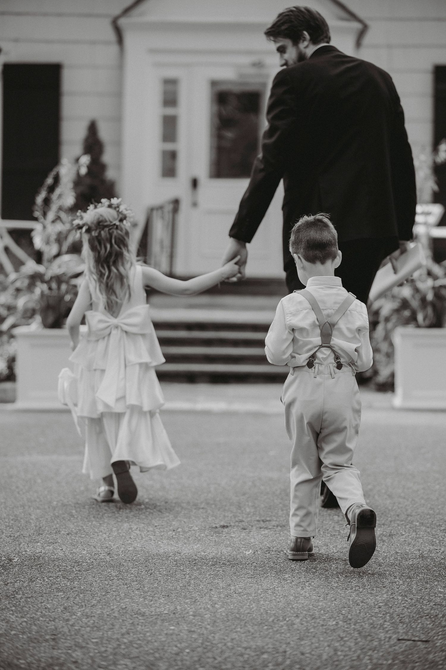 Black and white photo of groomsman walking flower girl and ring bearer toward a white wedding venue entrance.