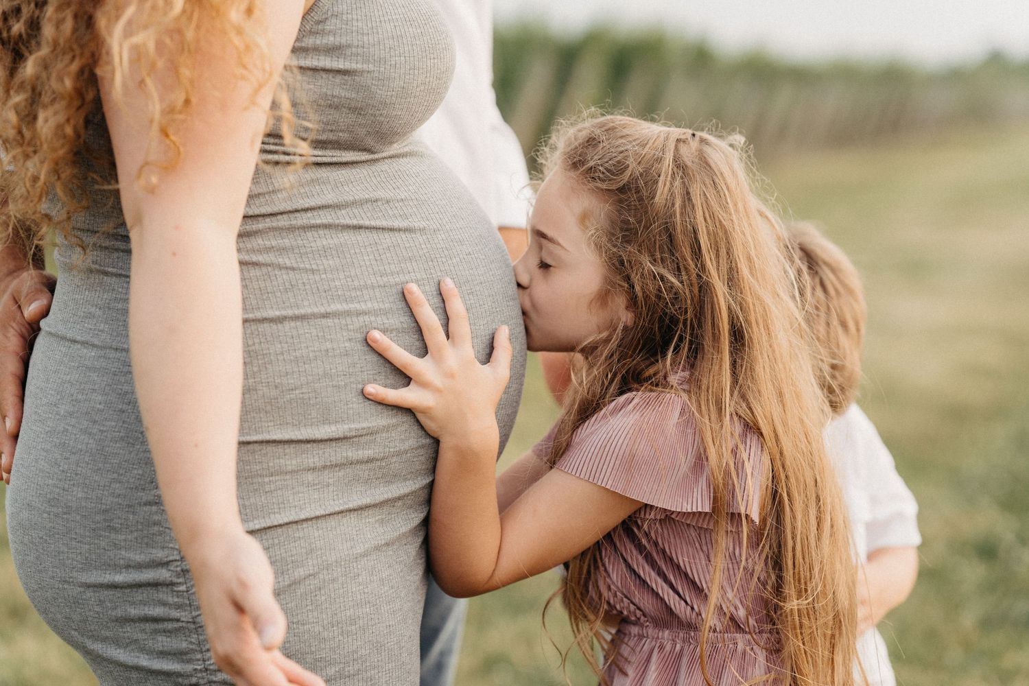 Young girl kissing pregnant mothers belly in field.