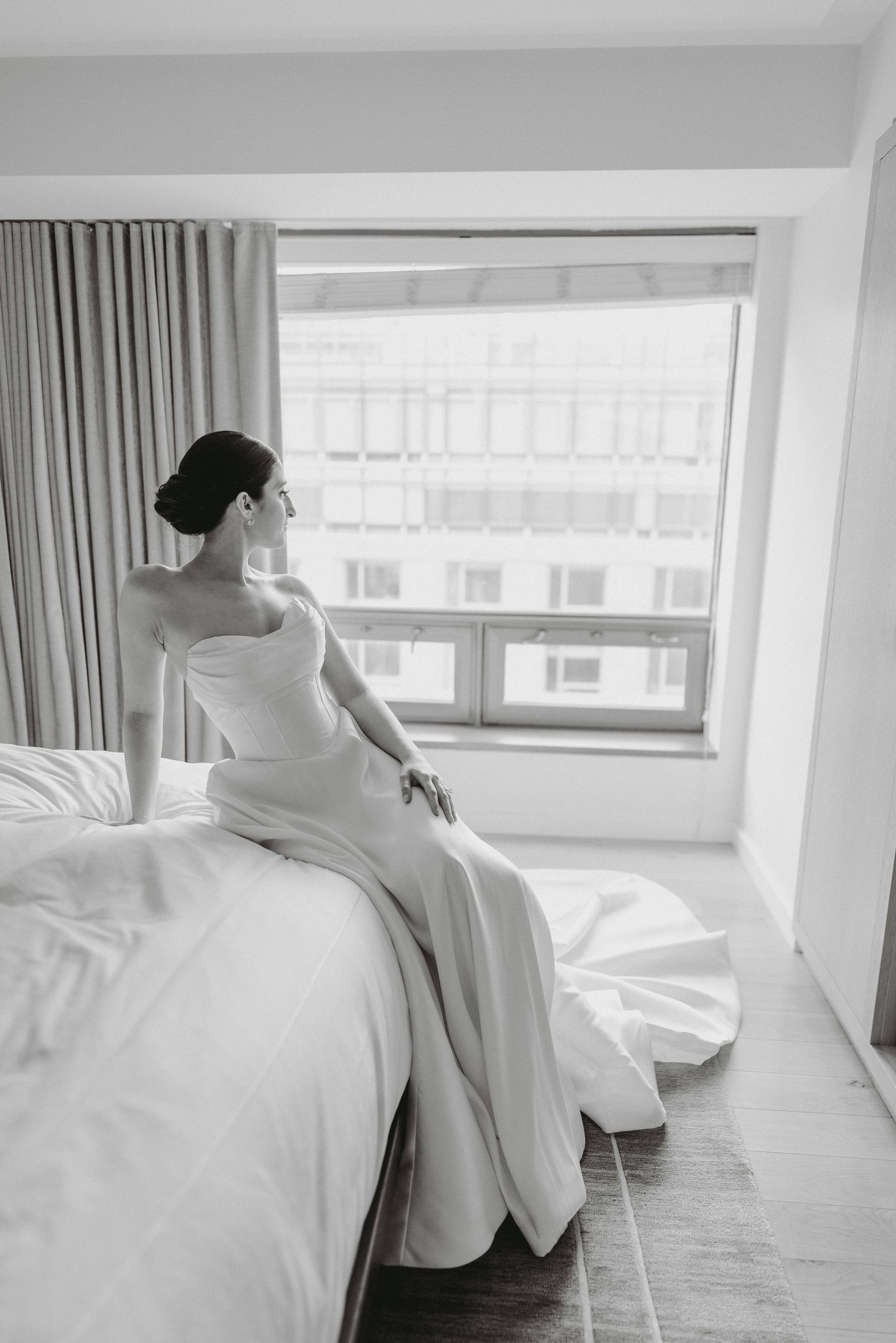 Black and white photo of bride in strapless gown sitting on a hotel bed gazing out the window.