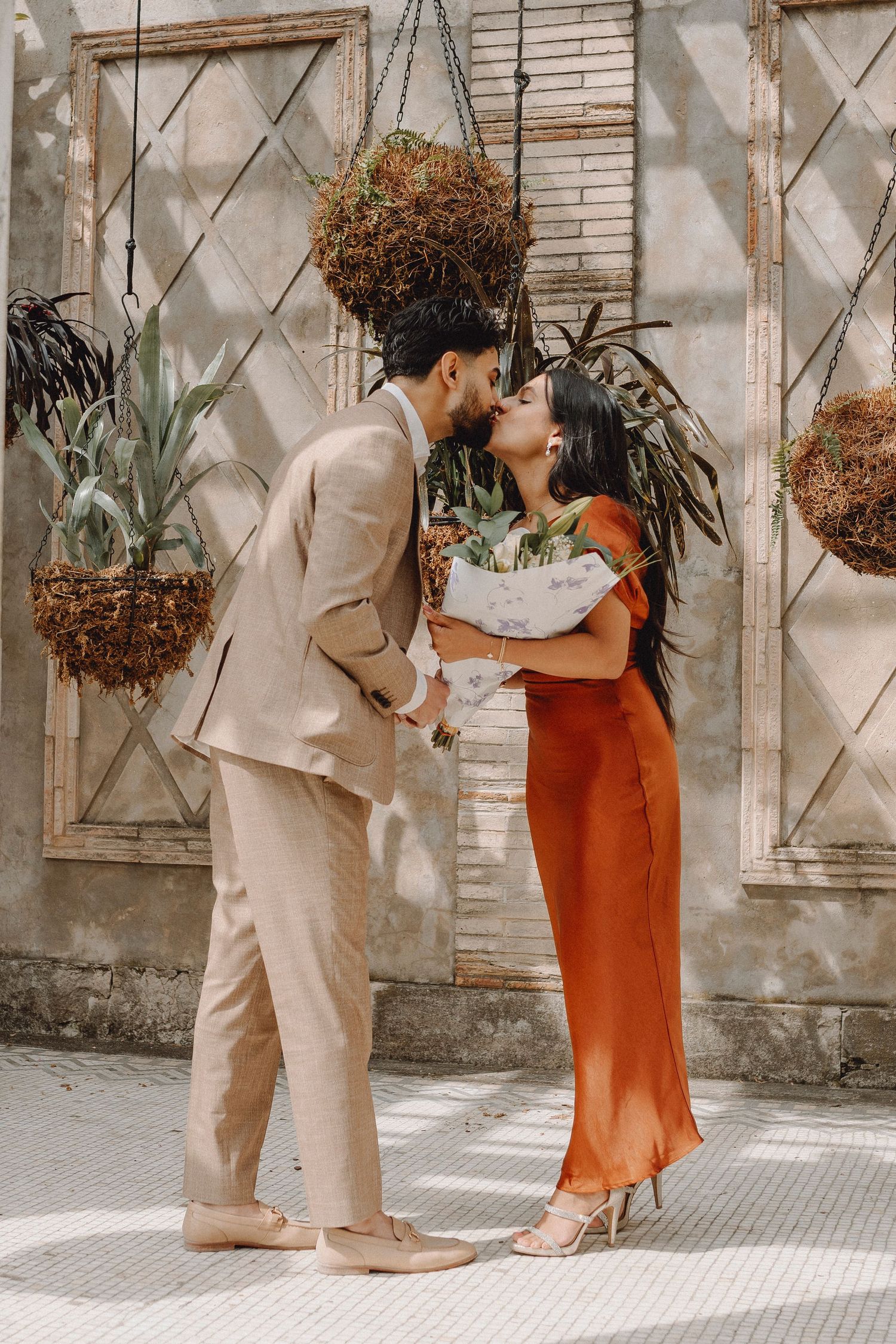 Couple shares a kiss while holding a floral bouquet in front of a rustic lattice wall.