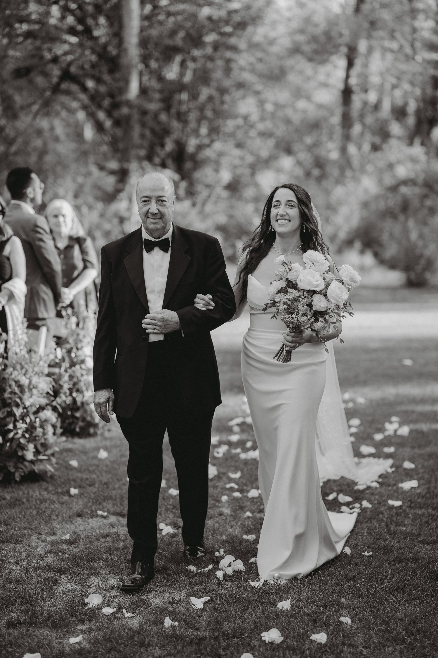 Black and white photo of bride walking down flower petal aisle with father during ceremony.