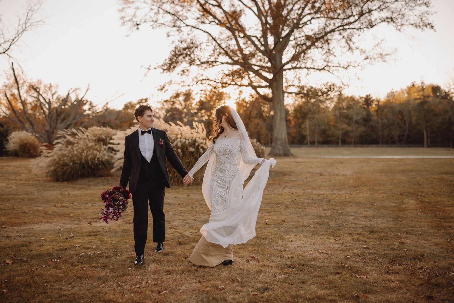 Bride and groom walking hand in hand across golden field at sunset in autumn park.