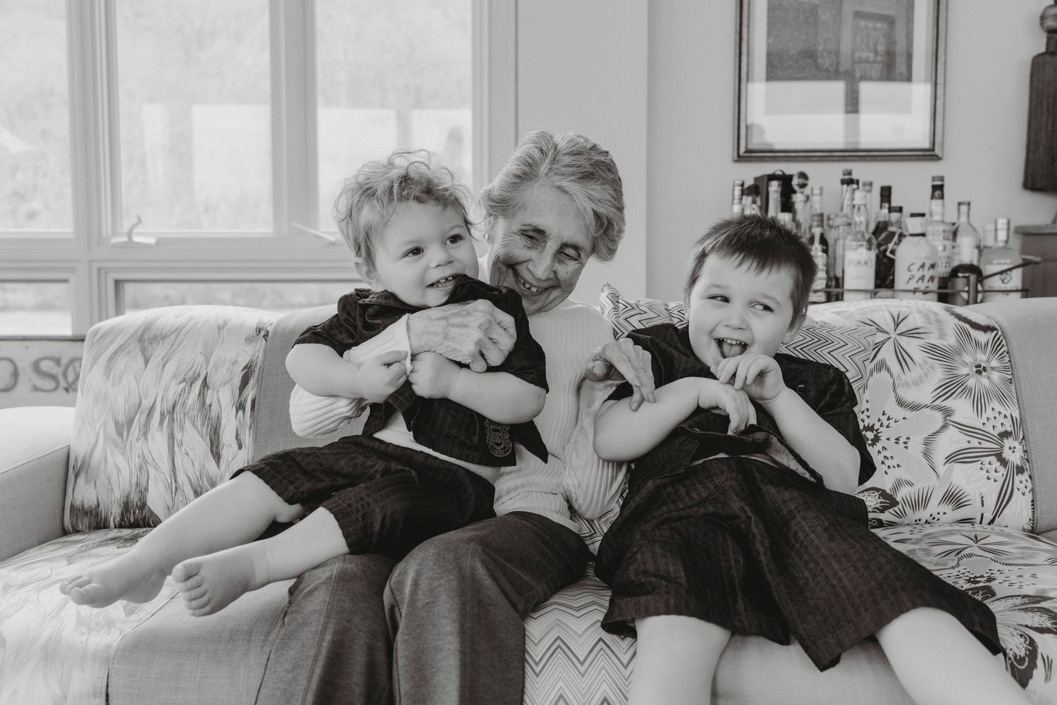 Grandmother laughing with two young children on a couch in a black and white family portrait.