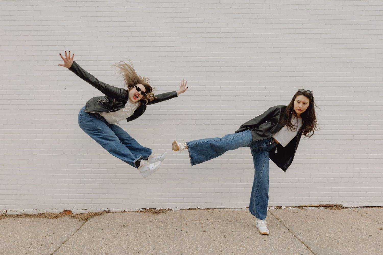Two young women in leather jackets and jeans strike dynamic poses against a white brick wall outdoors.