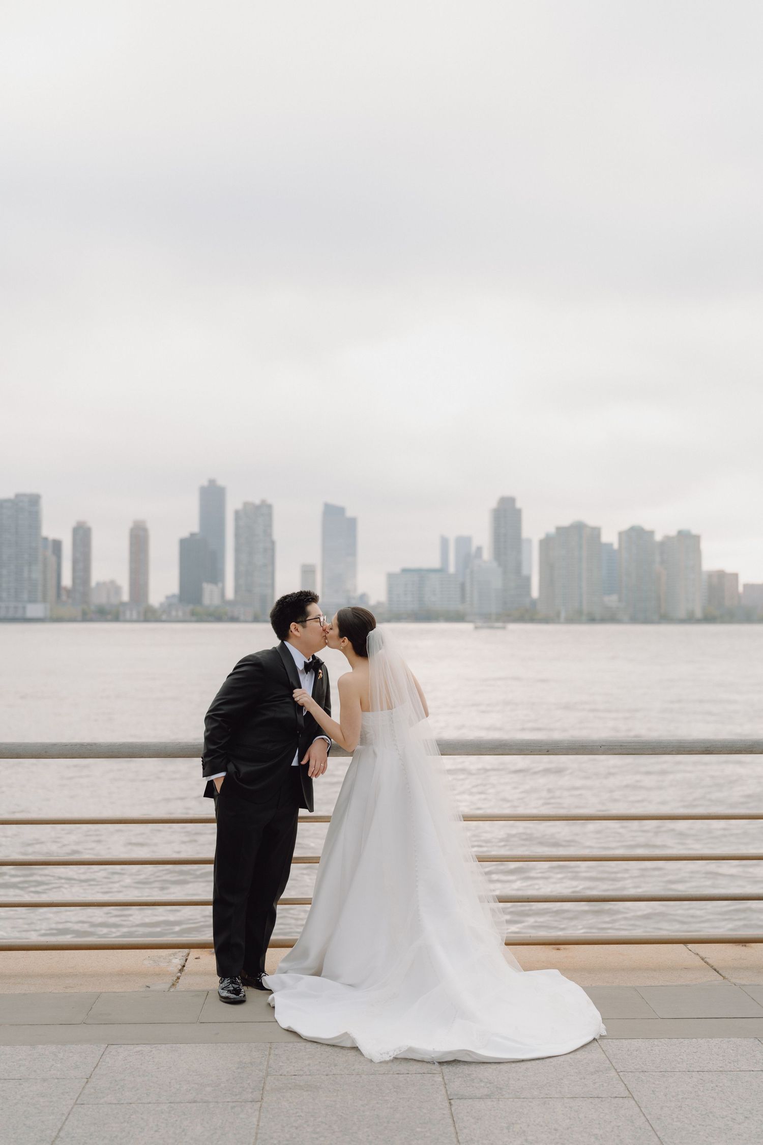 Bride and groom share a kiss on waterfront promenade with city skyline in the background.
