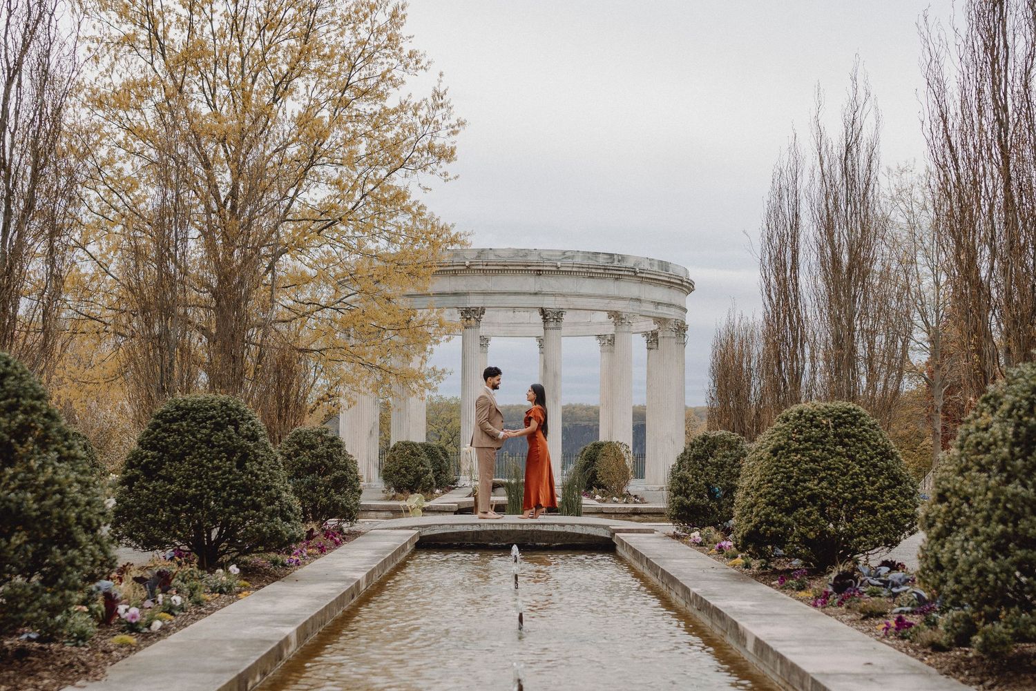 Couple holding hands by reflecting pool with classical columned rotunda backdrop and autumn trees in garden.