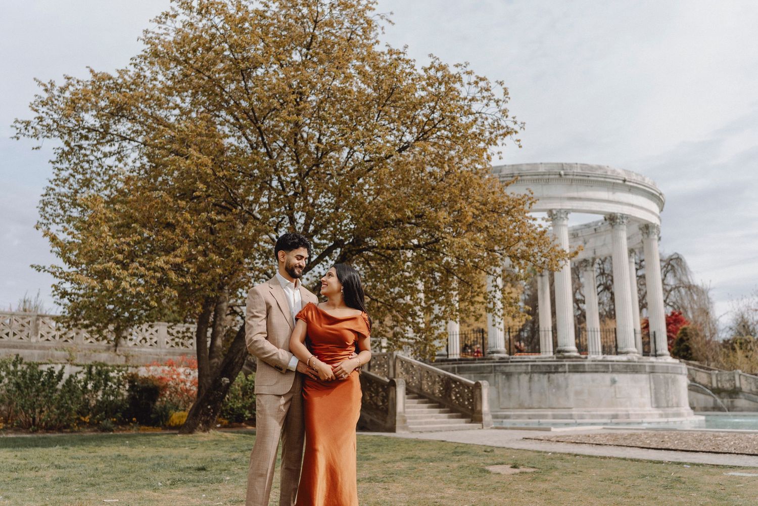 Couple in elegant attire standing before a white rotunda gazebo surrounded by autumn foliage.
