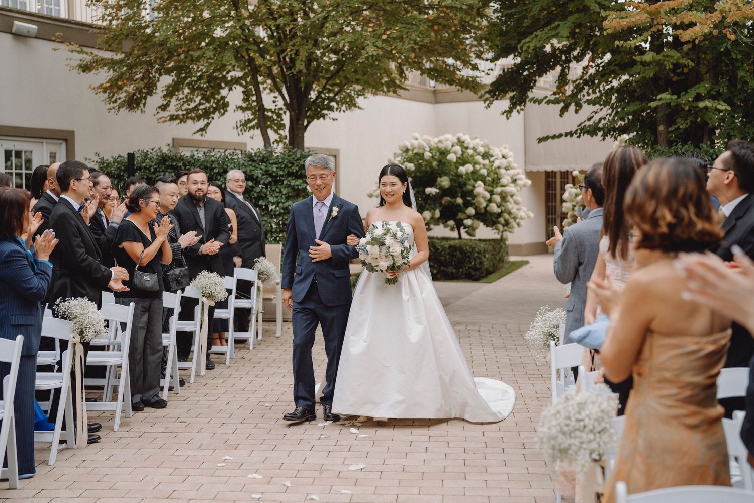 Father walks bride in strapless ball gown down outdoor aisle as wedding guests celebrate and applaud.