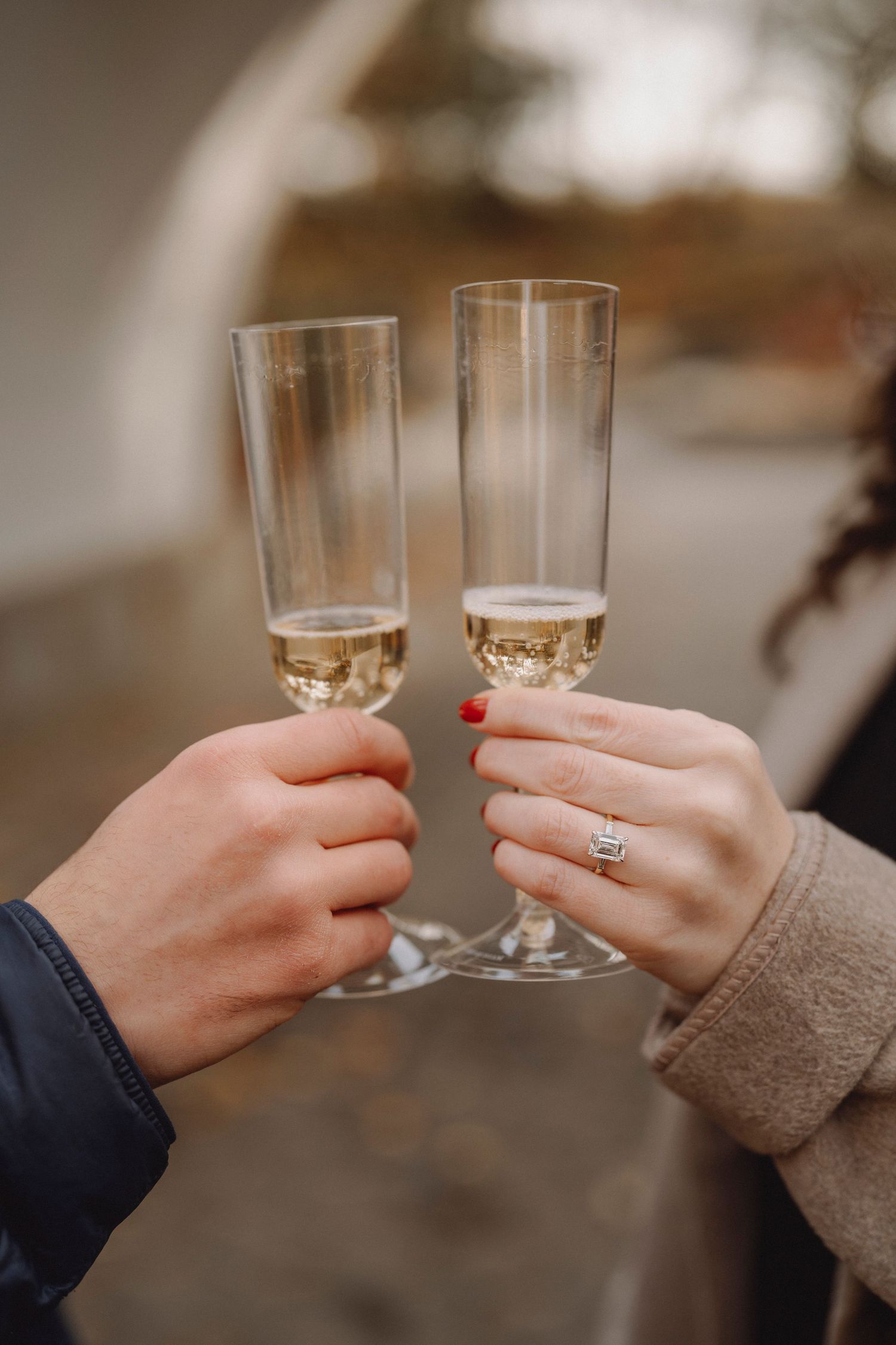Newly engaged couple clinking champagne flutes, woman showing diamond engagement ring with red nails.