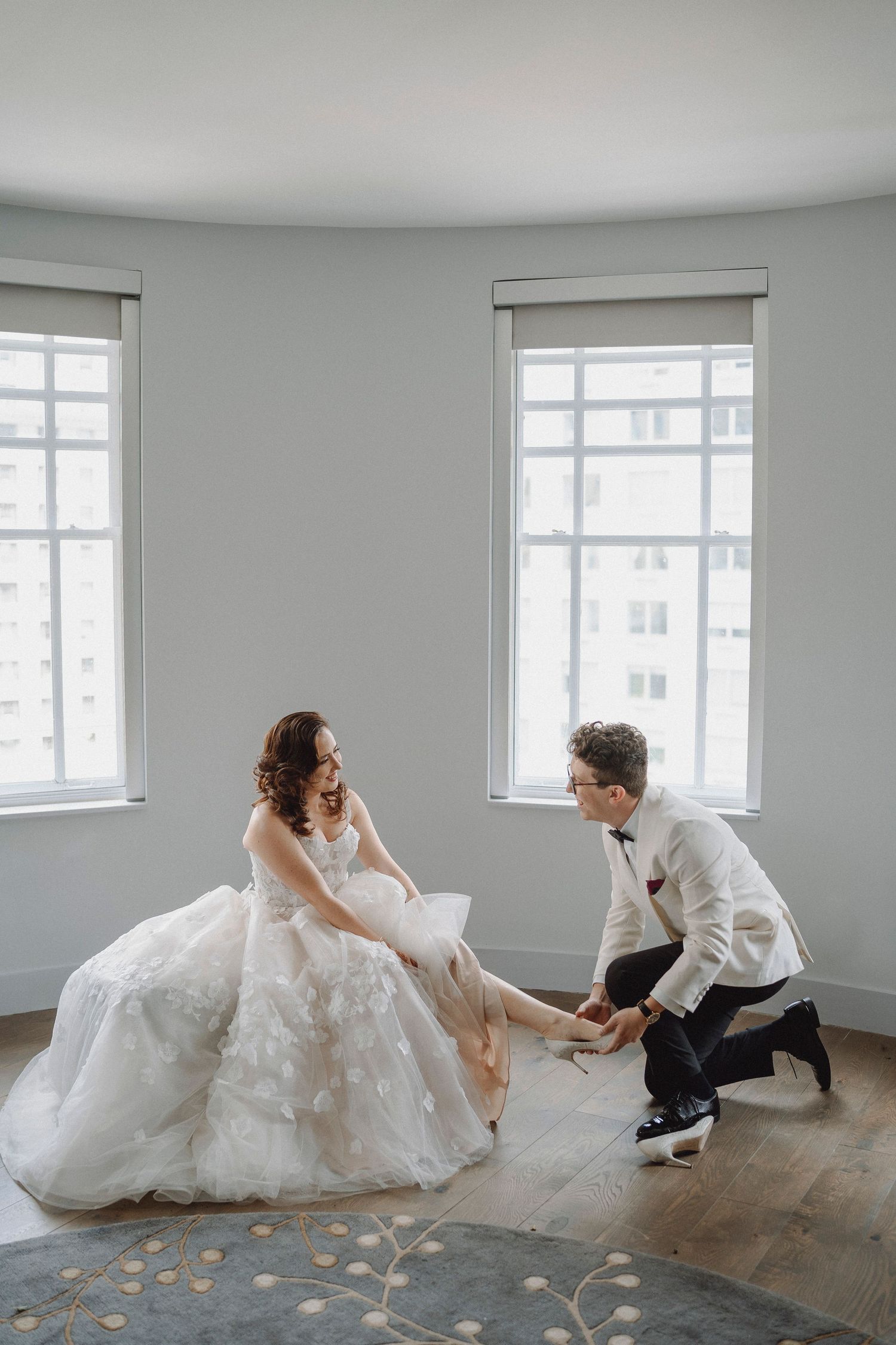 Groom in white tuxedo placing shoes on seated bride in elegant bridal suite.