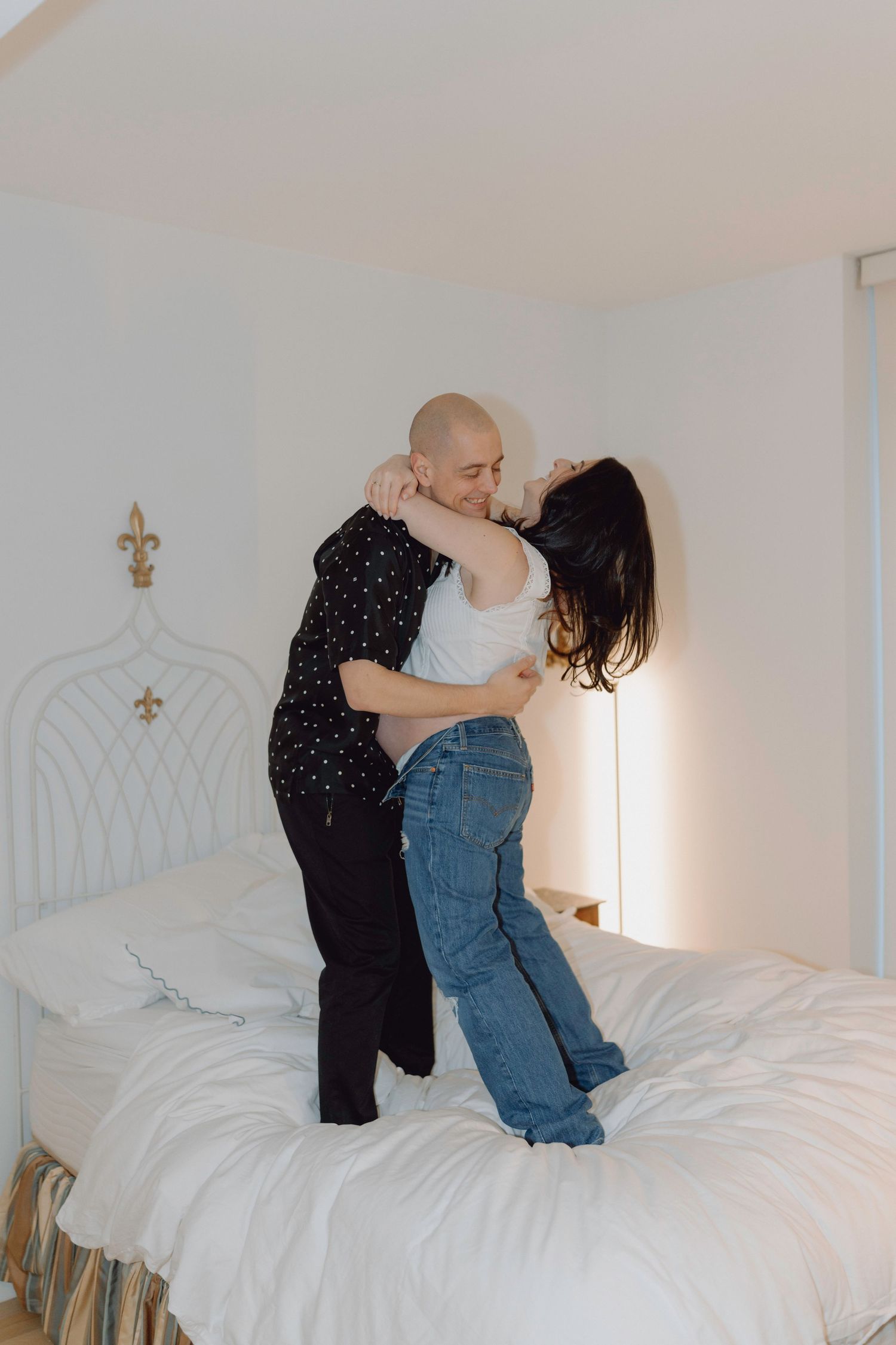 Couple kissing while standing on white bed in bright bedroom.