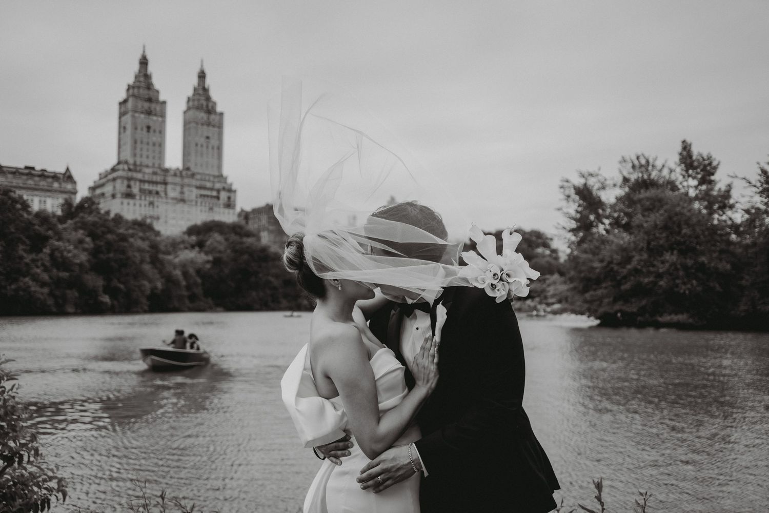 Black and white photo of couple kissing by Central Park lake with twin towers backdrop.