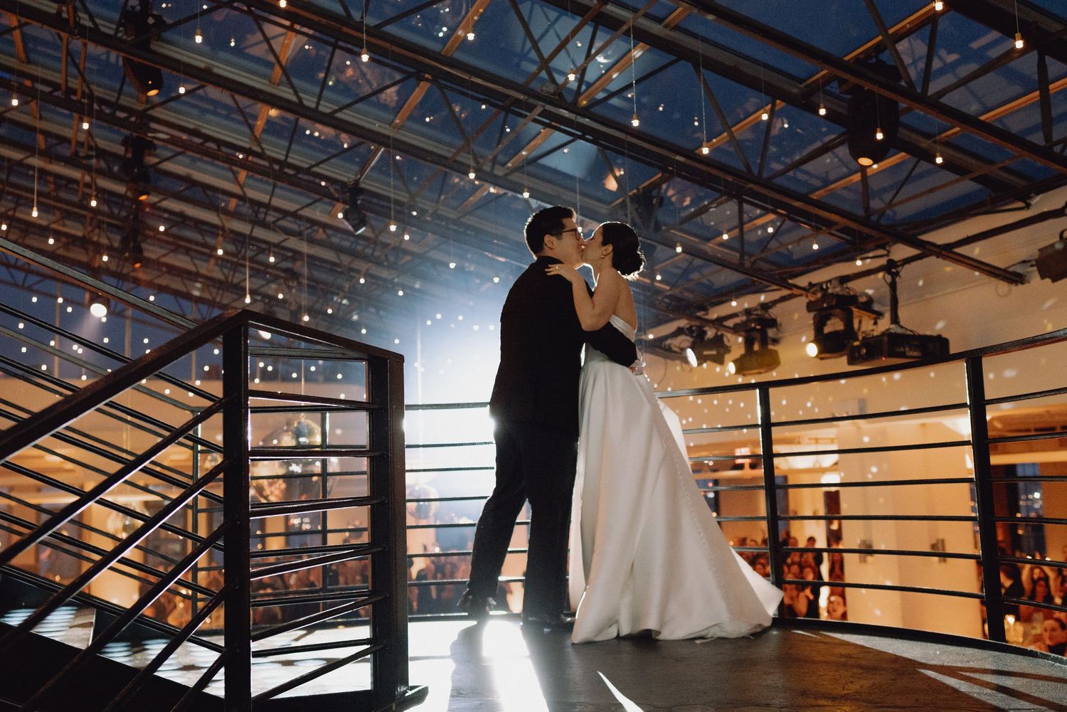 Bride and groom share romantic kiss on elevated platform in warmly lit industrial wedding venue.