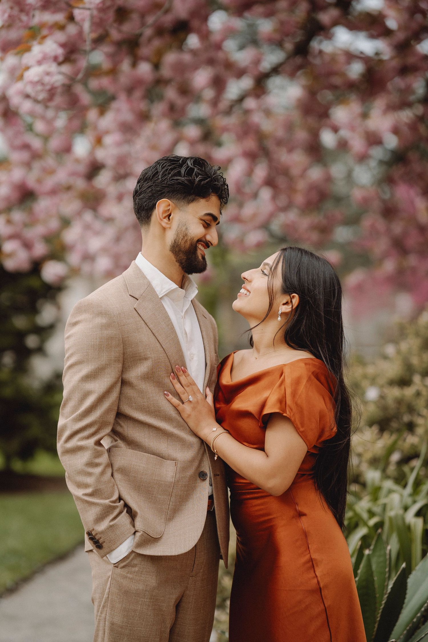 Couple gazing at each other laughing under pink cherry blossom trees, woman in rust dress, man in beige suit.