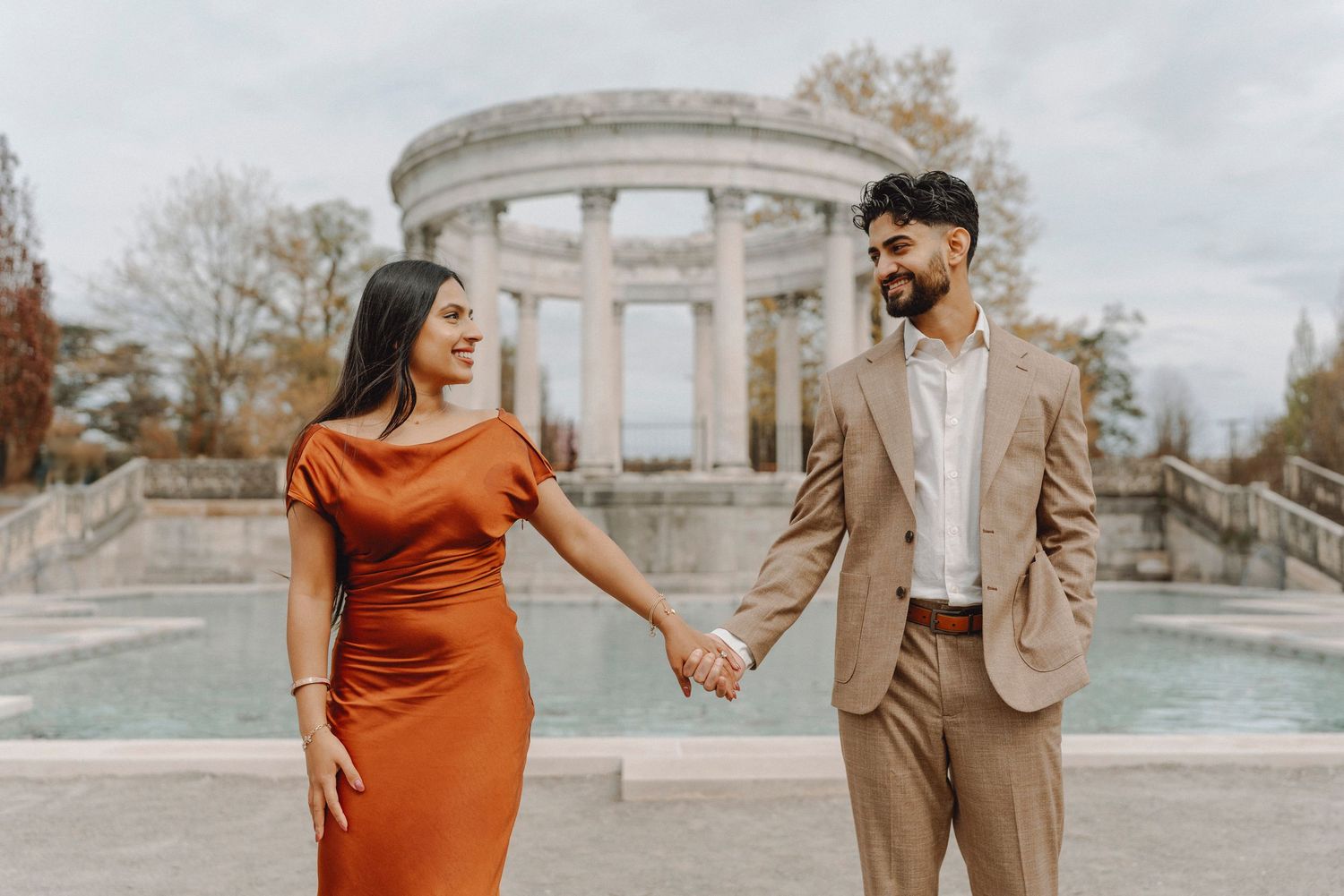 Smiling couple holding hands near a reflective pool with a neoclassical colonnade backdrop.