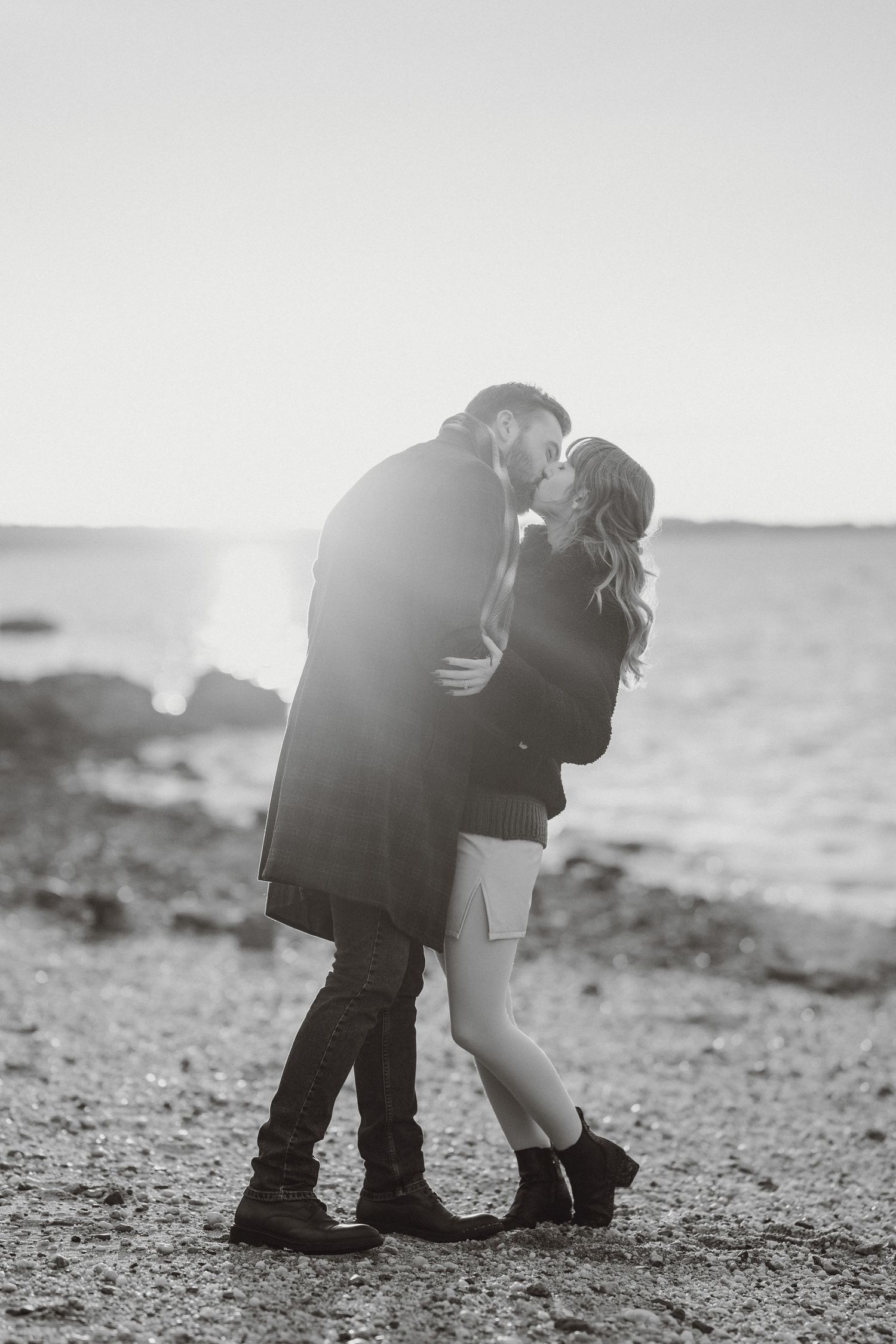 Couple sharing a romantic kiss on a sunlit rocky beach in dramatic black and white photo.