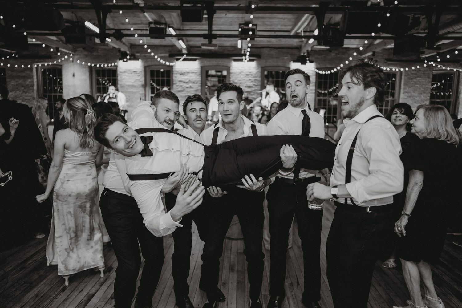 Groomsmen playfully lift groom horizontally at lively indoor wedding reception celebration.
