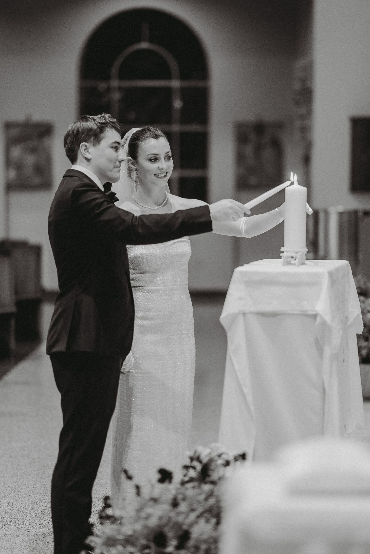 Black and white photo of smiling bride and groom lighting unity candle together during church wedding ceremony.