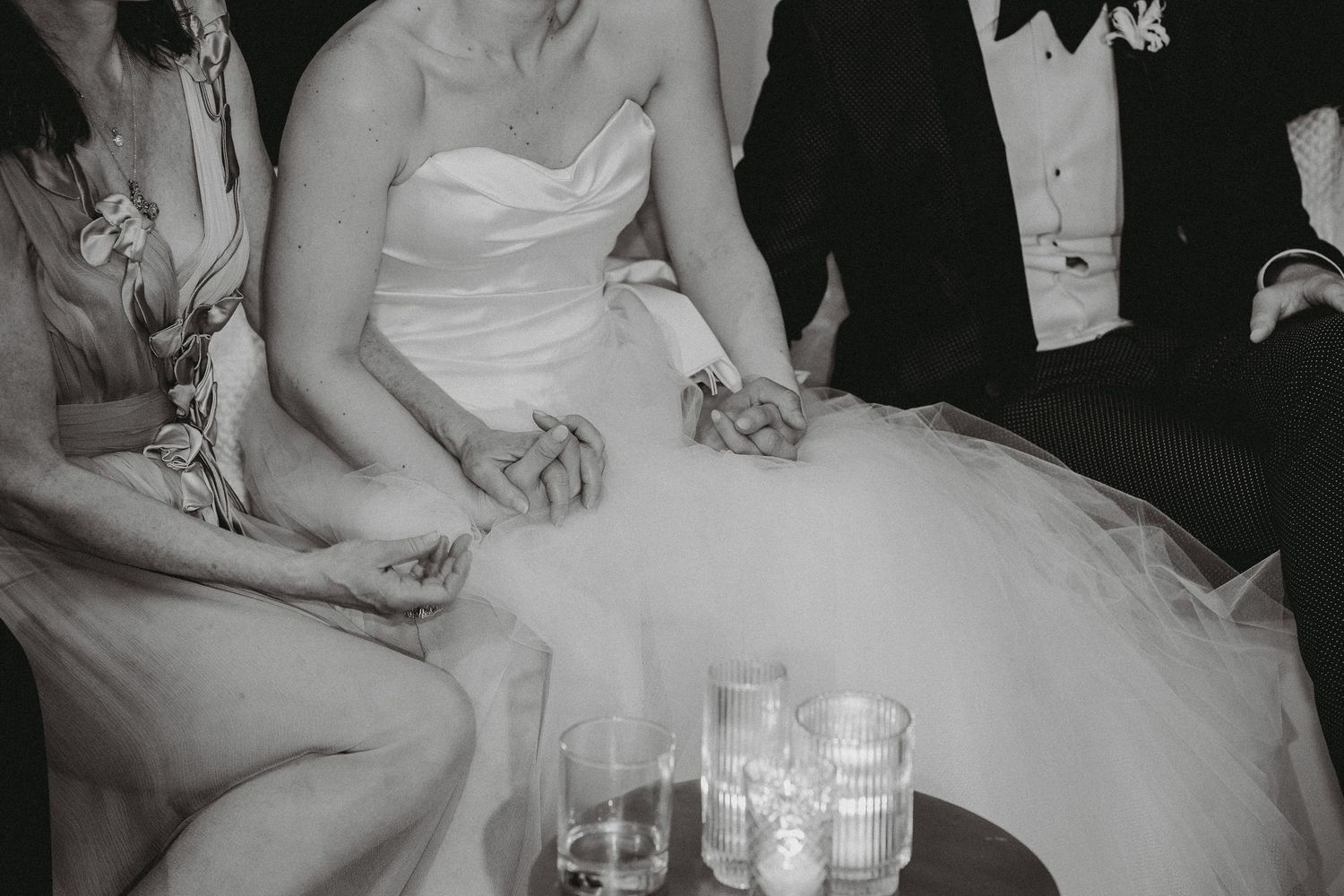 Black and white photo of bride holding hands with guests seated around candle-lit table at wedding reception.