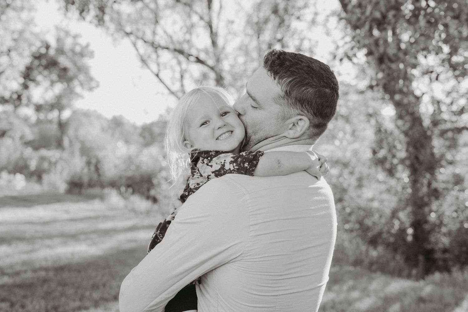 Father tenderly kissing his laughing young daughter in a black and white outdoor portrait.