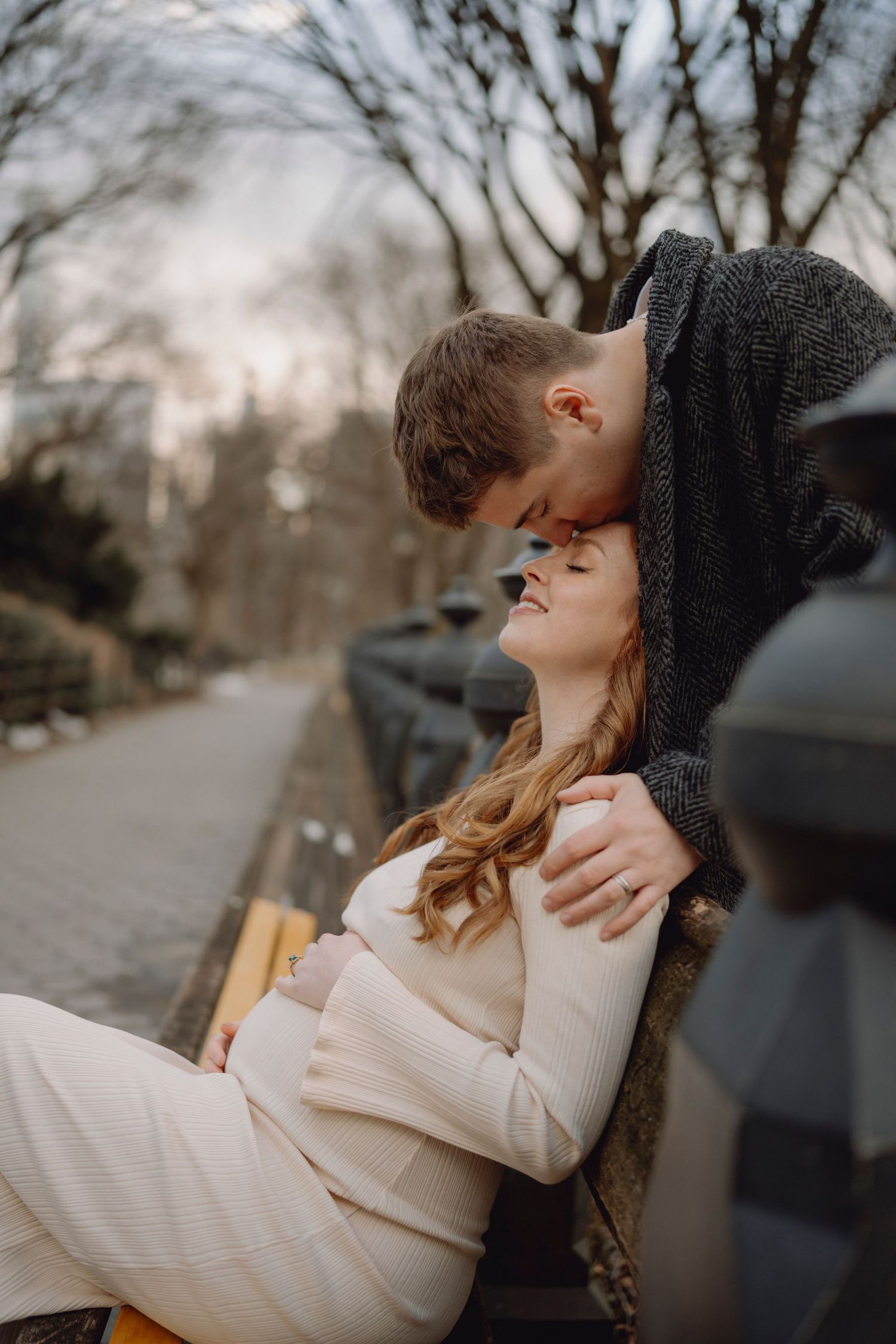 Tender moment as man kisses pregnant partners forehead on park bench at dusk.