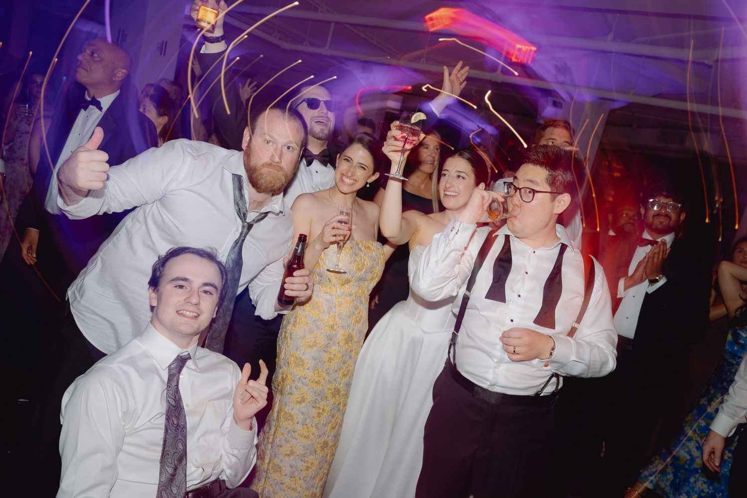 Group of wedding guests celebrating on dance floor, raising glasses and smiling at a festive reception party.