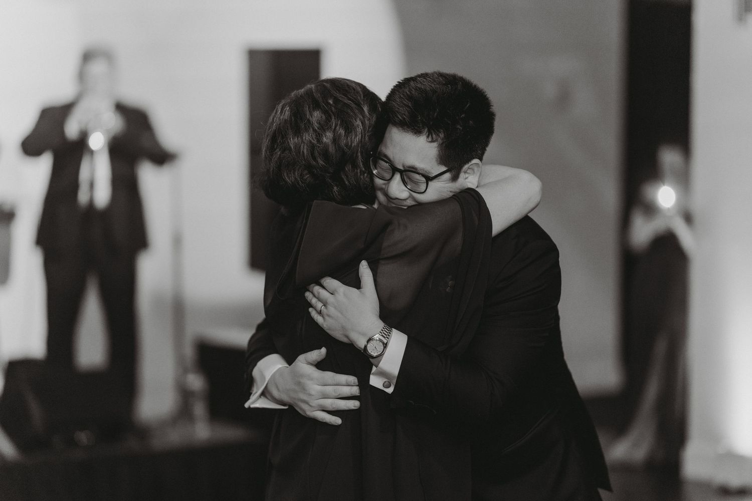 Emotional black and white photo of groom sharing a heartfelt hug with a guest at wedding reception.