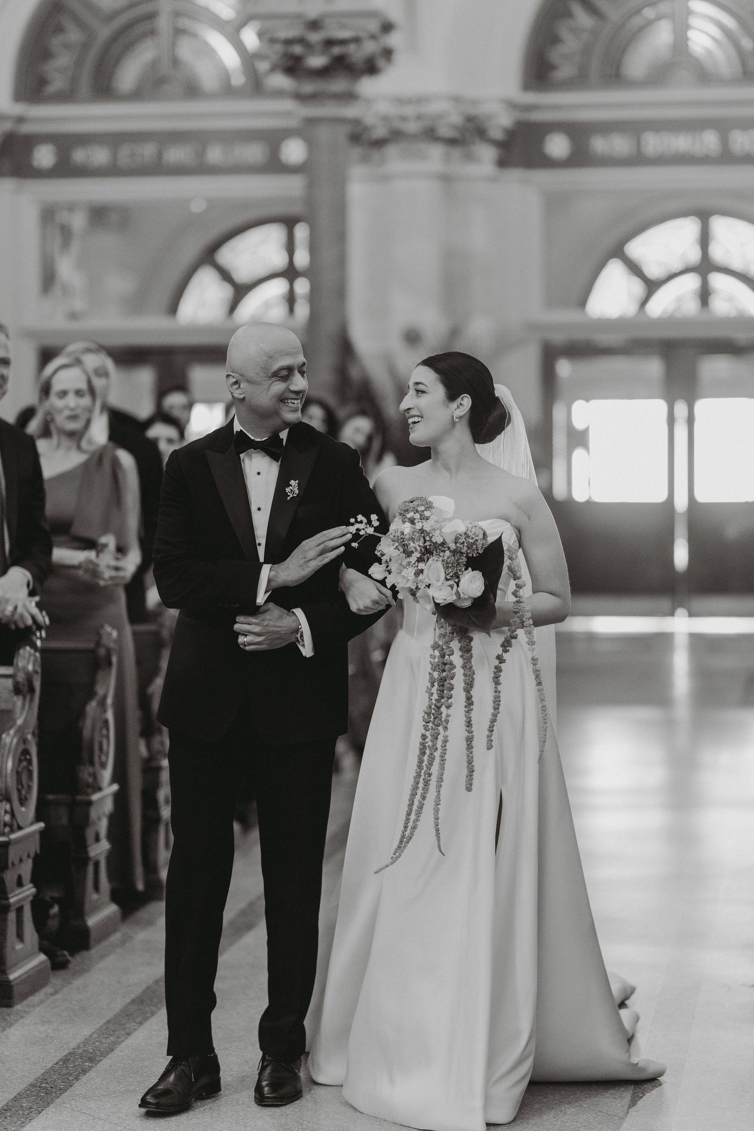 Black and white photo of bride and escort sharing joyful moment walking down church aisle.