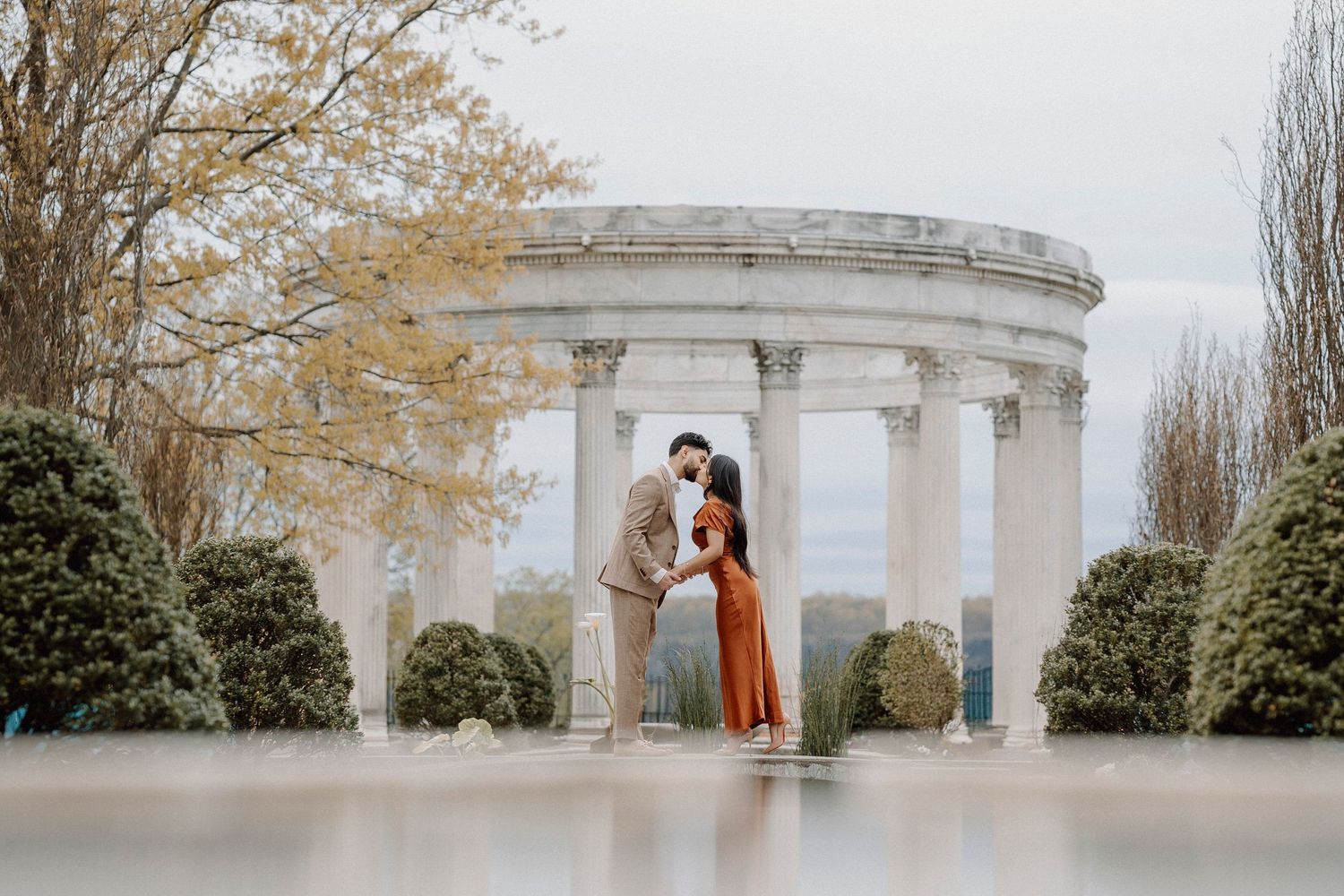 Couple kissing under classical stone rotunda with columns, surrounded by manicured gardens and misty landscape.