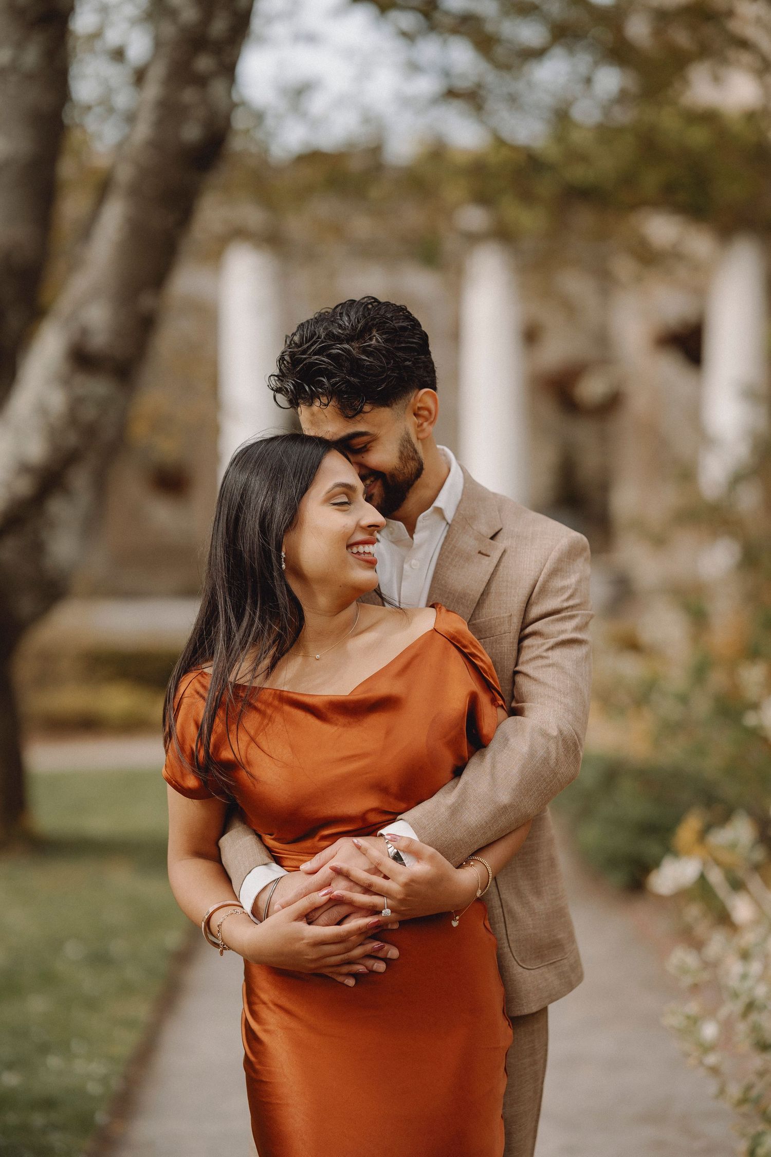 Couple embracing in a garden, woman in rust satin dress smiling as man in beige suit holds her from behind.