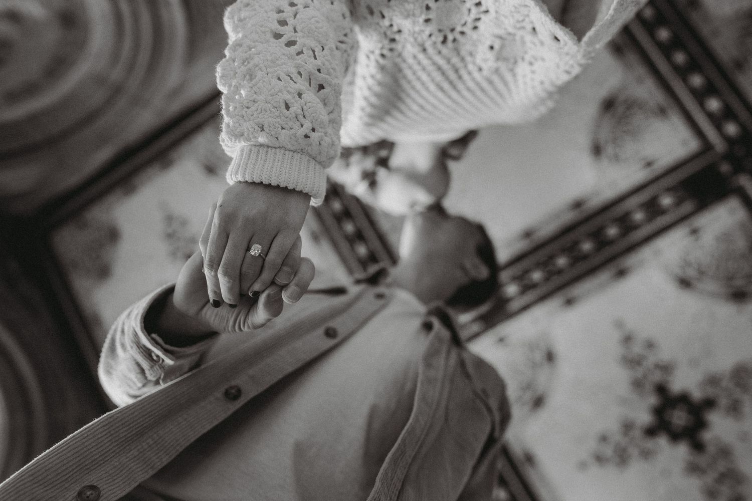 Black and white close-up of an engaged couple holding hands, showcasing a diamond engagement ring.
