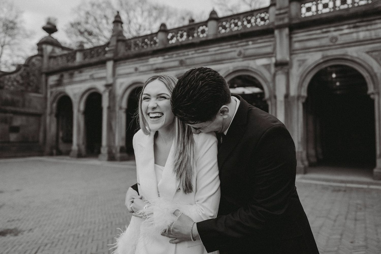Black and white photo of a bride laughing as groom nuzzles her outside Bethesda Terrace.