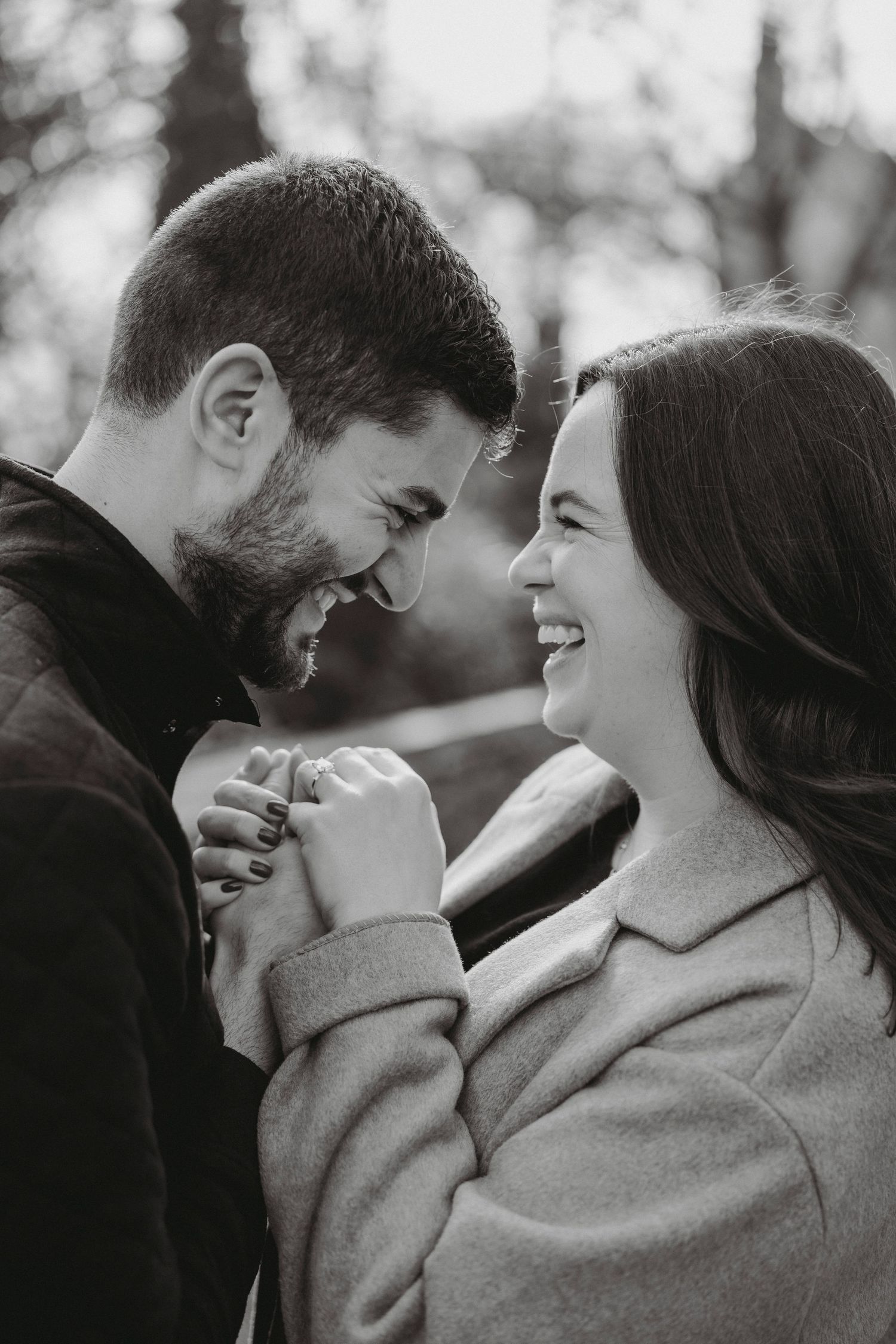 Black and white photo of joyful couple laughing and holding hands after marriage proposal in park.