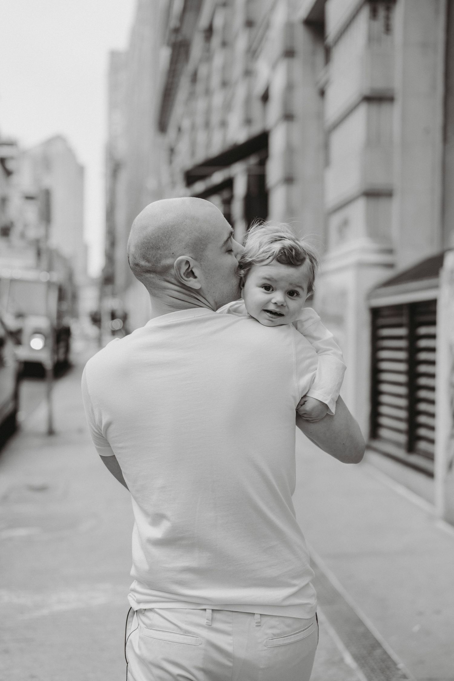 Bald father kissing his baby while holding them up on a city street in black and white.