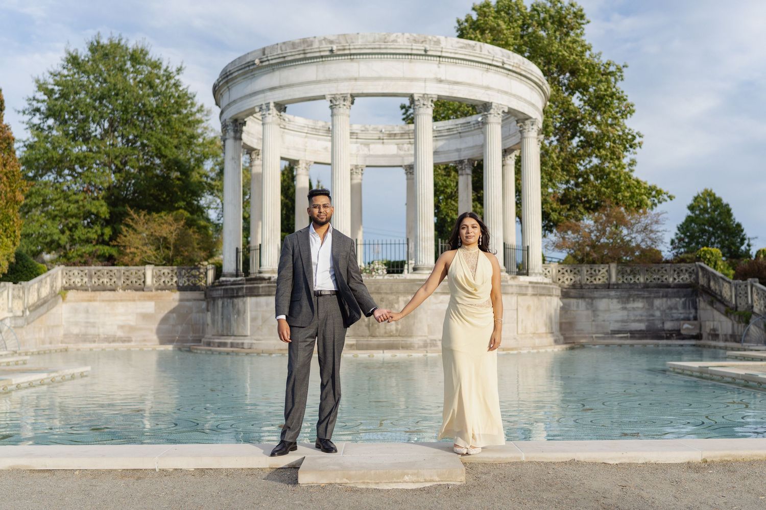 Couple holding hands in front of a white columned neoclassical pavilion and reflecting pool, man in a gray suit and woman in a cream gown.
