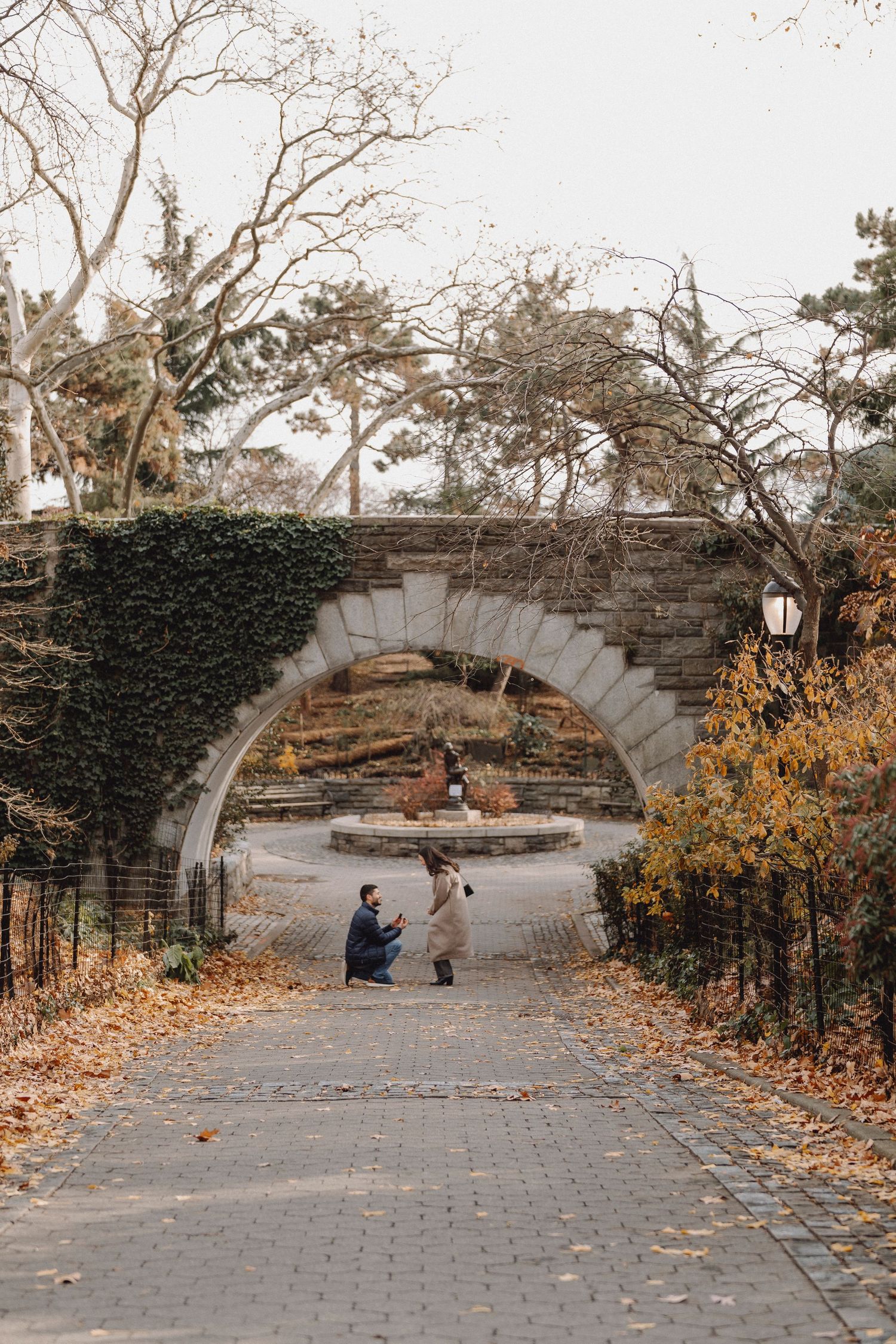 Man kneeling to propose to a woman on a brick path beneath a vine-covered stone arch in a park, surrounded by colorful autumn foliage.