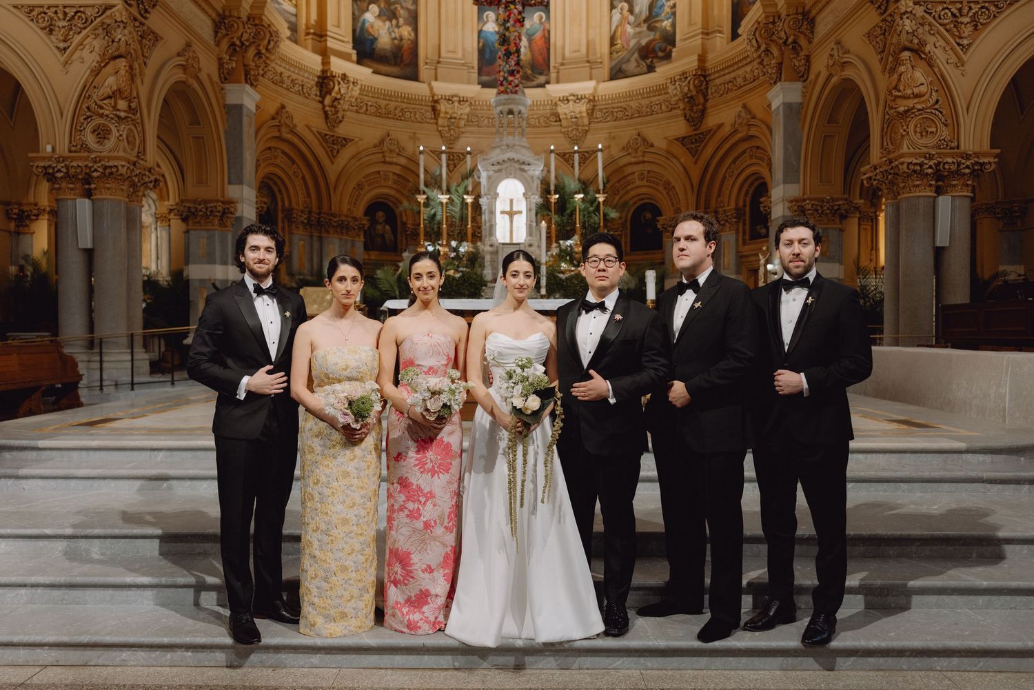 Wedding party poses on church altar steps, bridesmaids in floral gowns and groomsmen in black tuxedos.