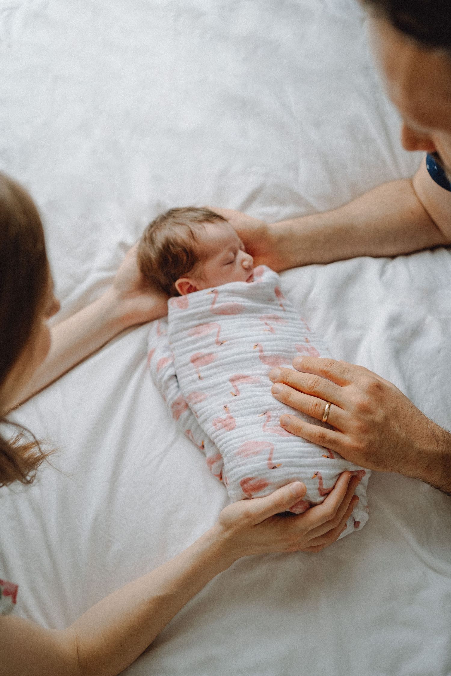 New parents gently touching swaddled sleeping newborn baby on white bed.