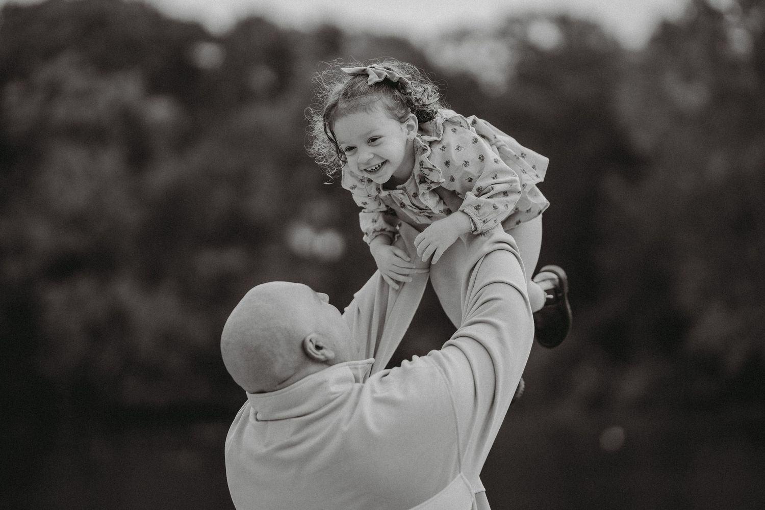 Black and white photo of a bald father lifting his laughing toddler daughter high in the air outside.