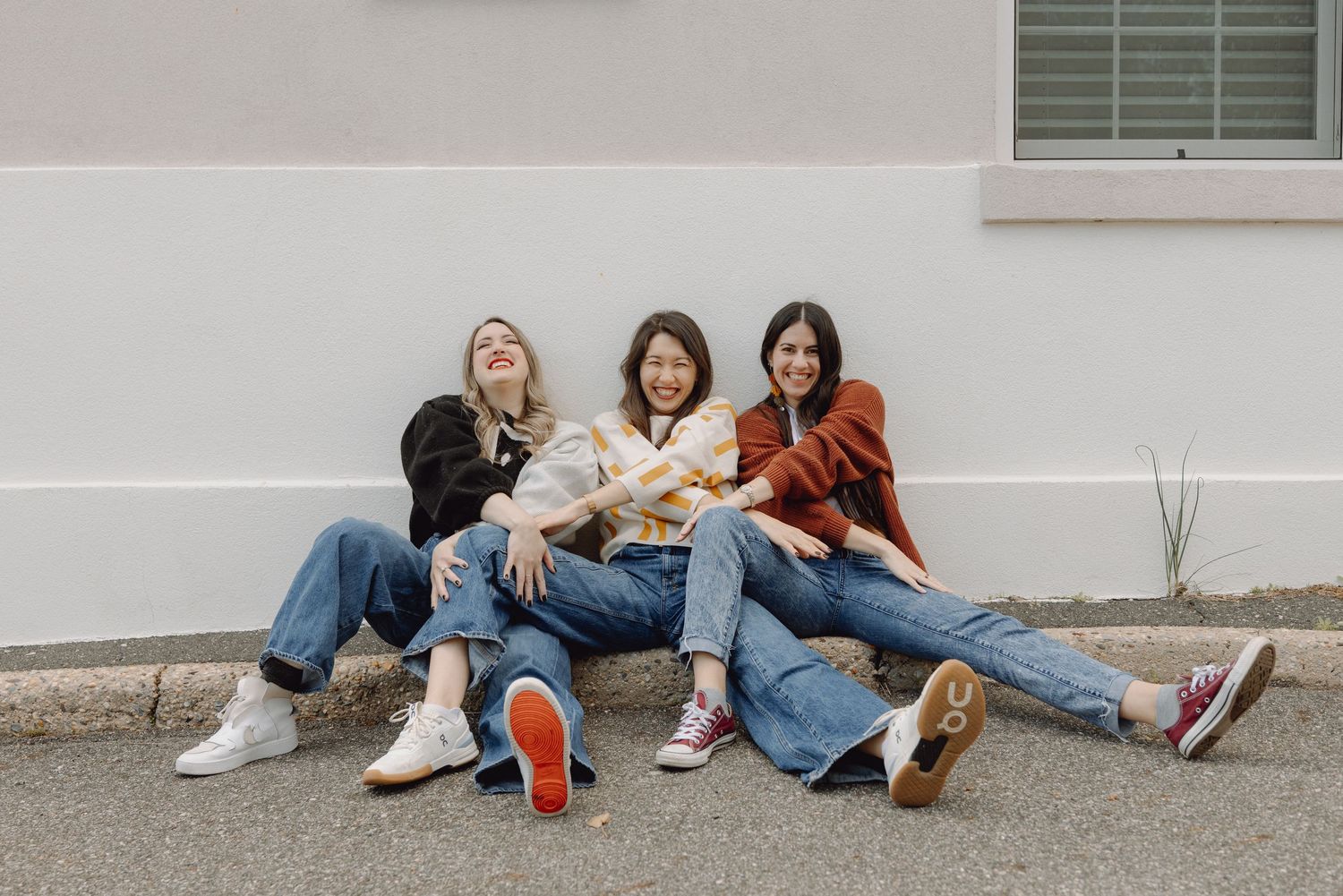 Three young women in wide-leg jeans sit laughing together against a white wall outdoors.