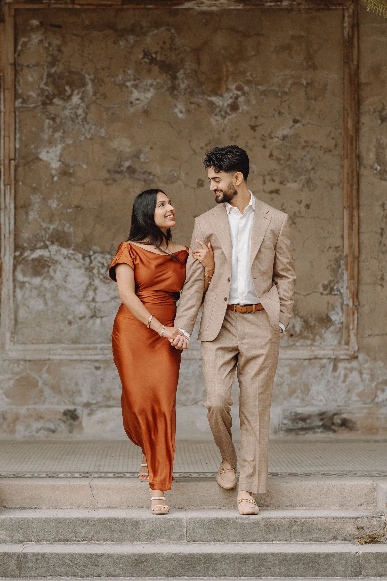Couple holding hands and smiling at each other while descending stone steps by a rustic wall.