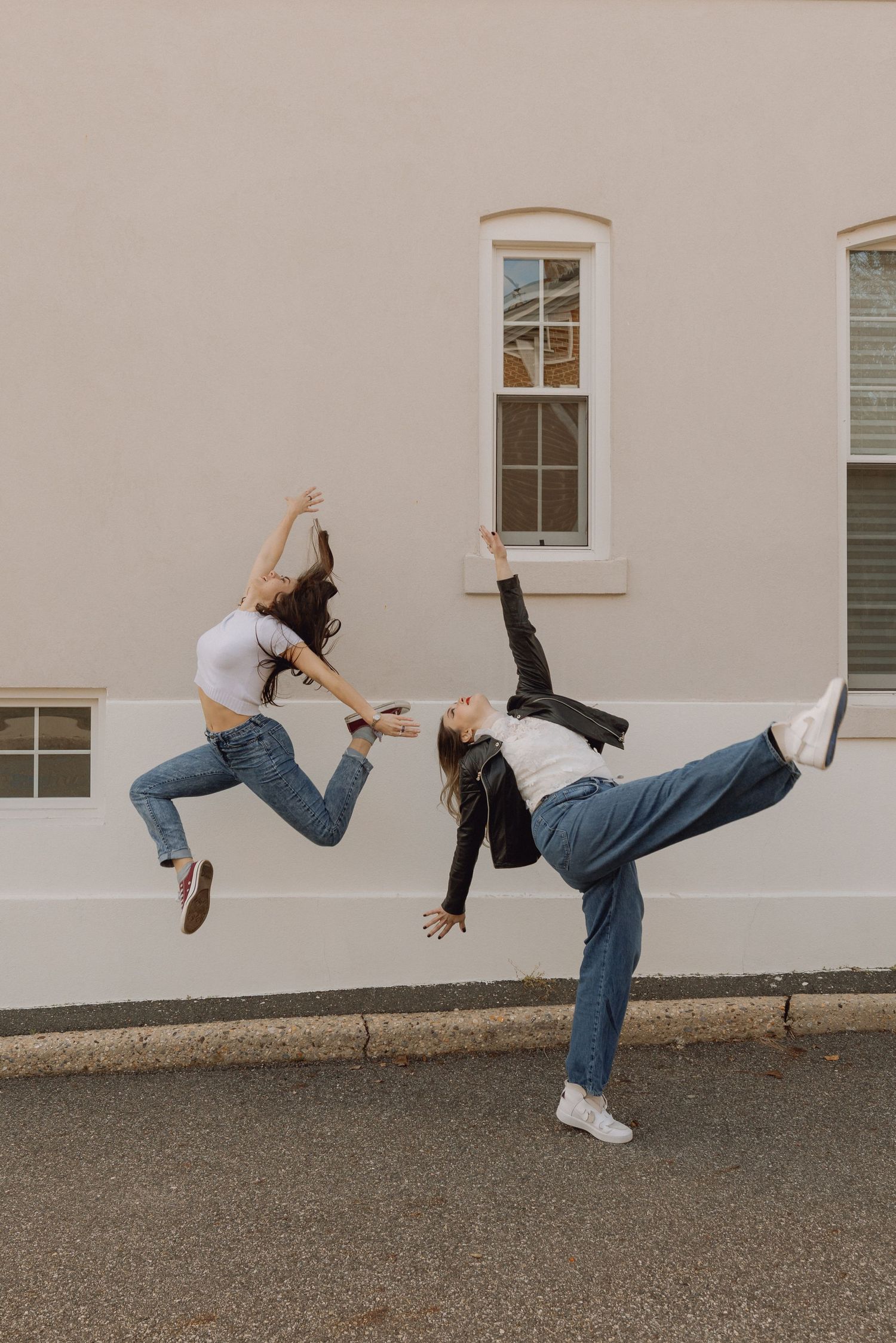 Two young women jumping and dancing energetically in front of a beige building wearing jeans and casual tops.